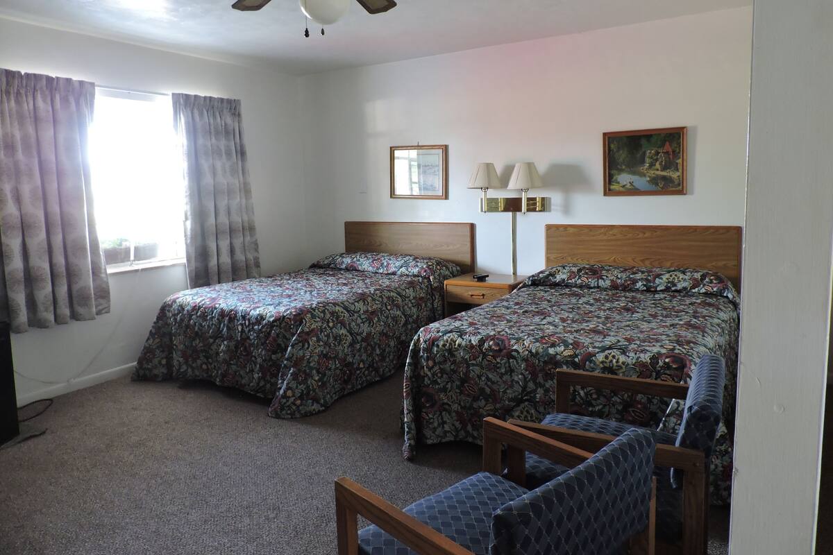 Habitación de hotel con dos camas, un ventilador de techo, una mesita de noche con lámpara y un cuadro en la pared.