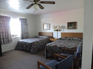 A hotel room with two beds, a ceiling fan, a nightstand with a lamp, and a painting on the wall.