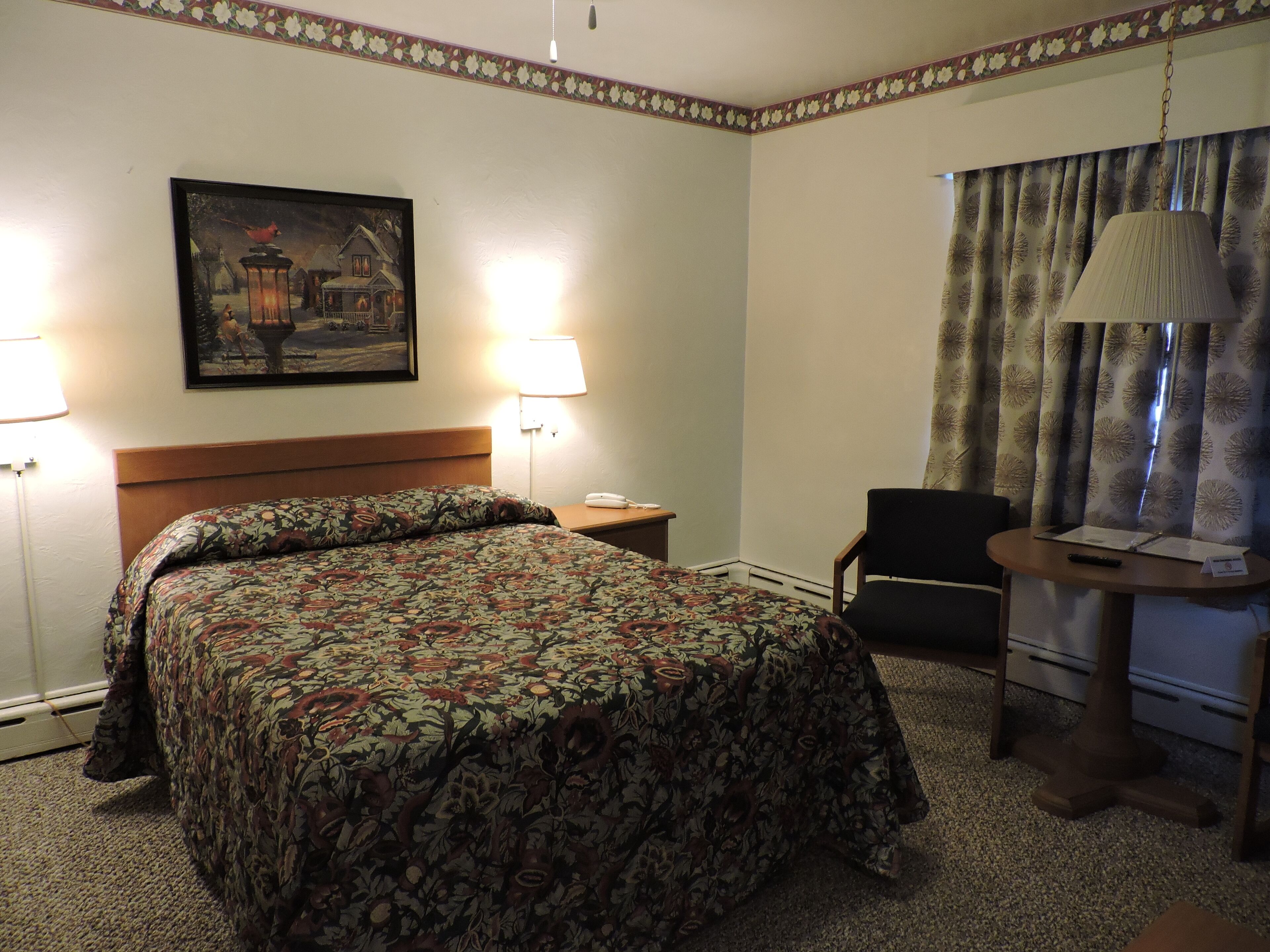 A hotel room with a bed, two bedside lamps, a desk with a lamp, a chair, and a painting on the wall.