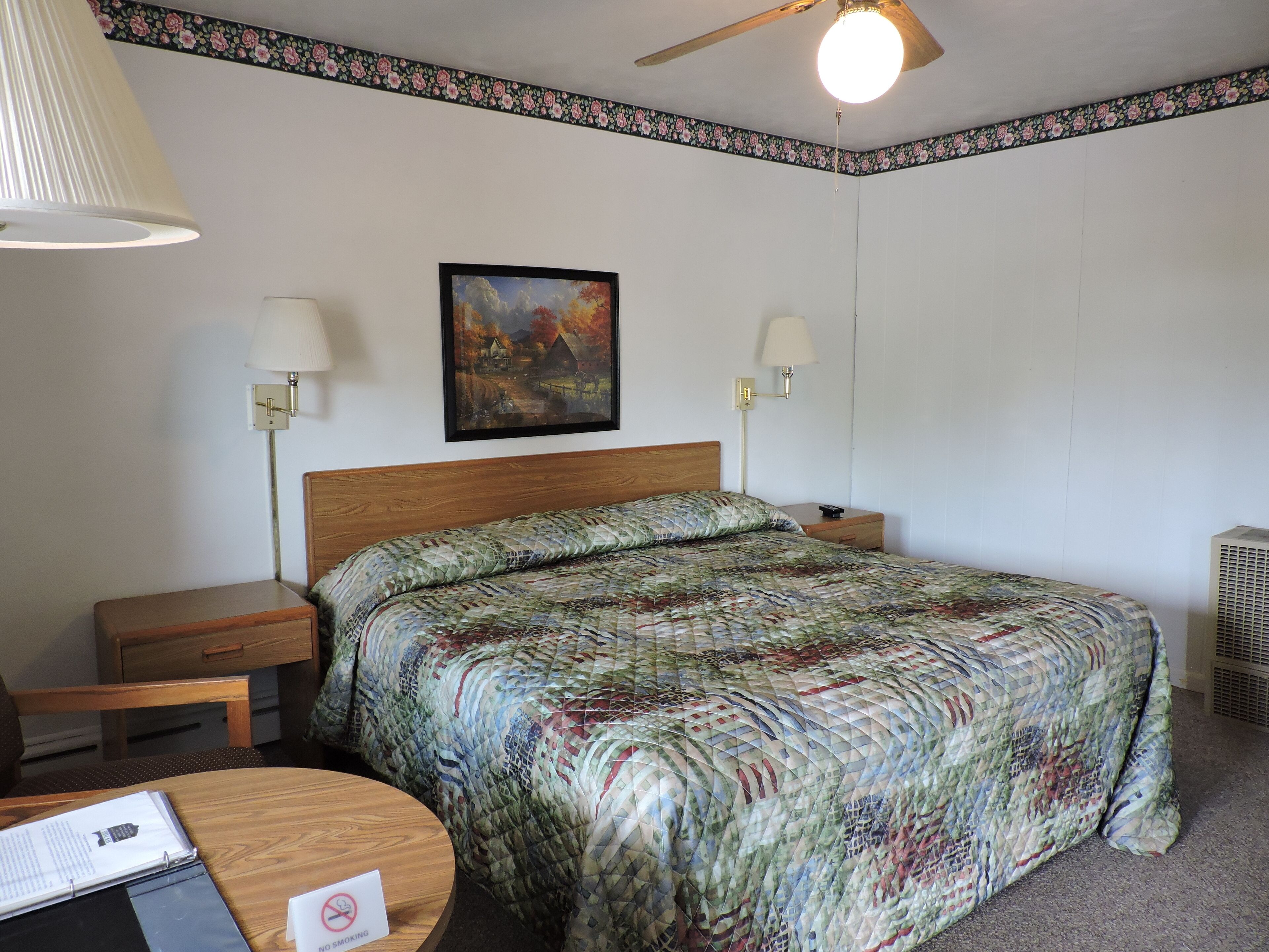 A hotel room with a wooden bed, two bedside lamps, a painting on the wall, a desk with a chair, and a no-smoking sign.