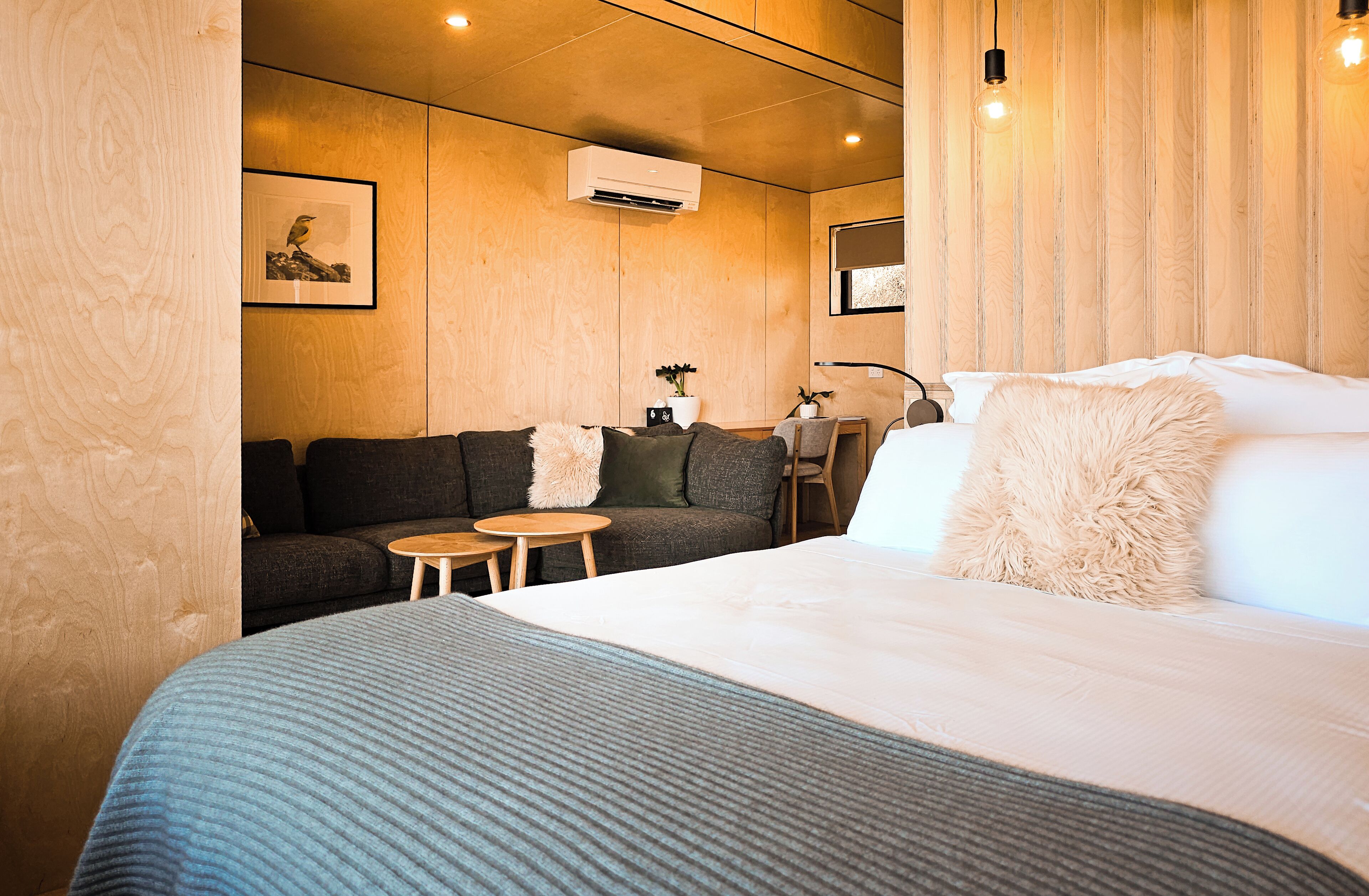 Quarto de hotel moderno com uma cama grande, um sofá, duas mesinhas e um ar condicionado fixado na parede.