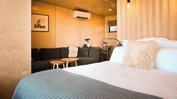 Quarto de hotel moderno com uma cama grande, um sofá, duas mesinhas e um ar condicionado fixado na parede.