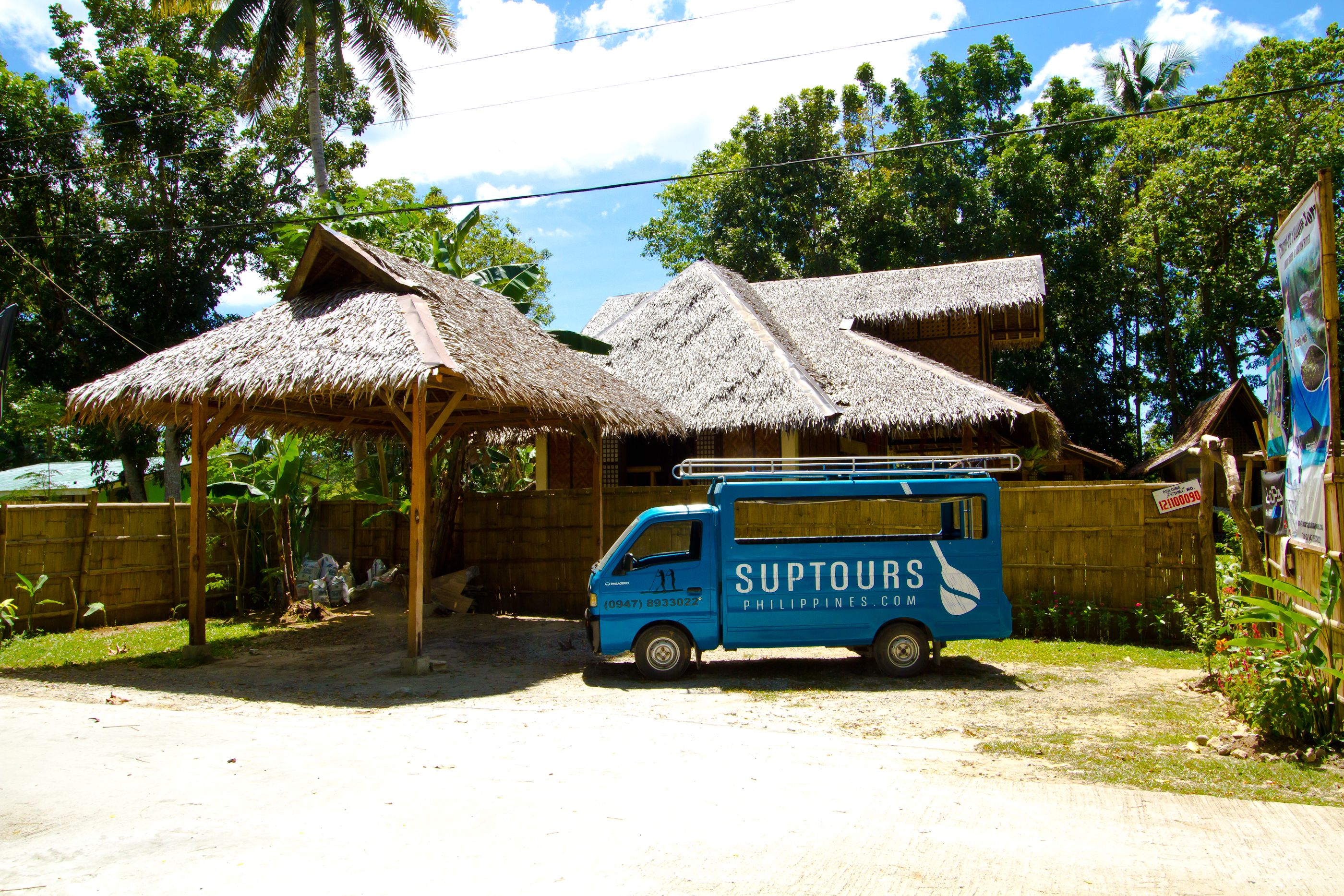 Una camioneta azul con la leyenda "SUPTOURS PHILIPPINES.COM" estacionada frente a una casa con techo de paja.