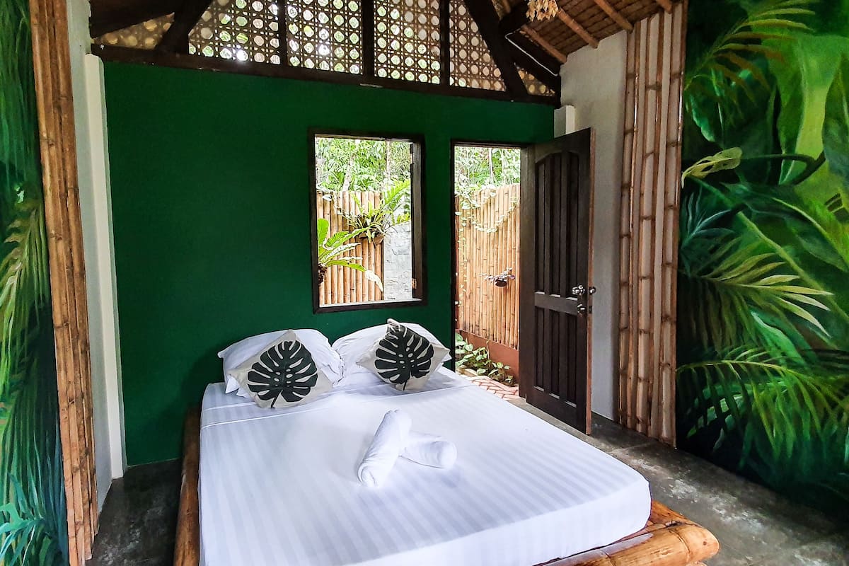 Una cama con colchón blanco y ropa de cama a rayas, flanqueada por paredes verdes y decoraciones con motivos de hojas.