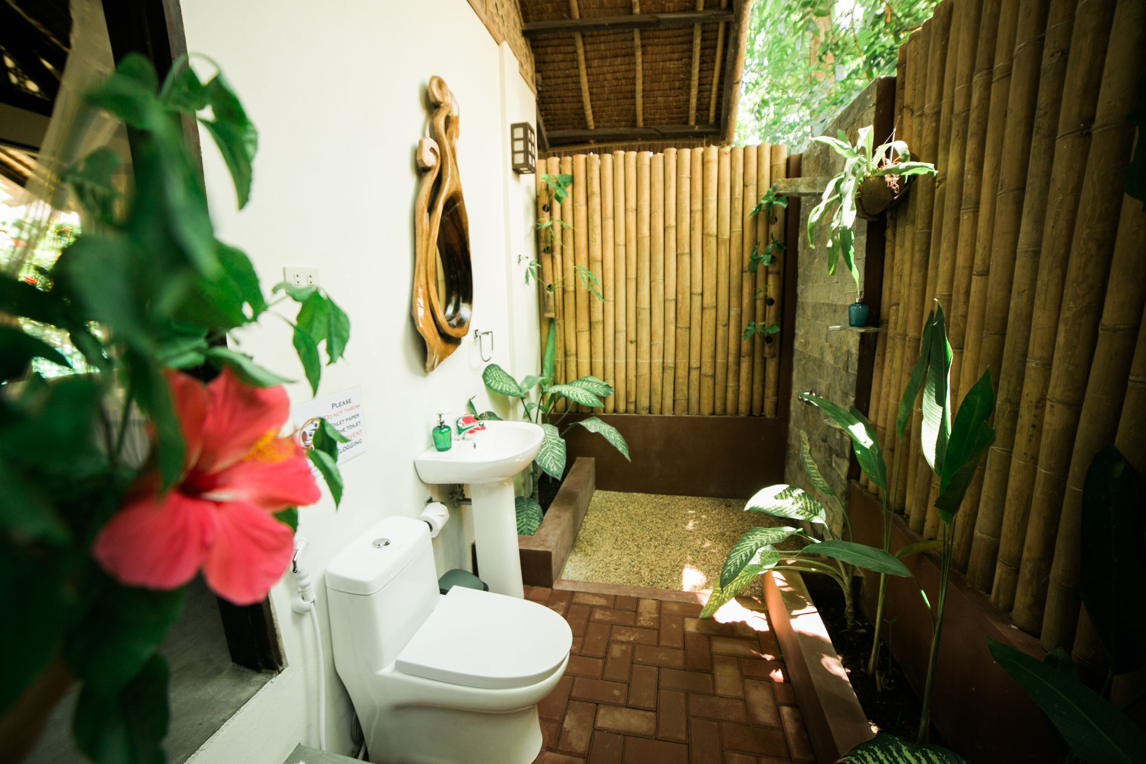 A compact bathroom with a toilet, sink, and shower area, surrounded by bamboo walls and potted plants.
