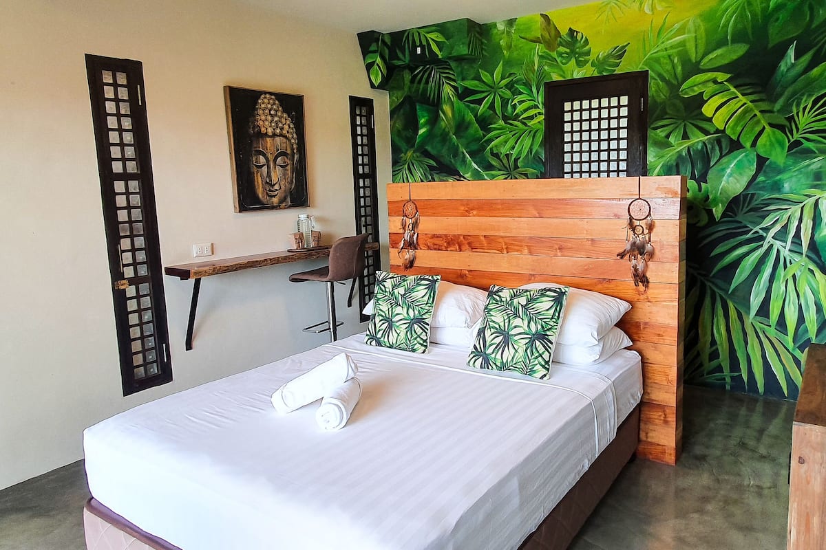 Un dormitorio con una cama, cabecera de madera, un mural en la pared, una mesita y una silla.