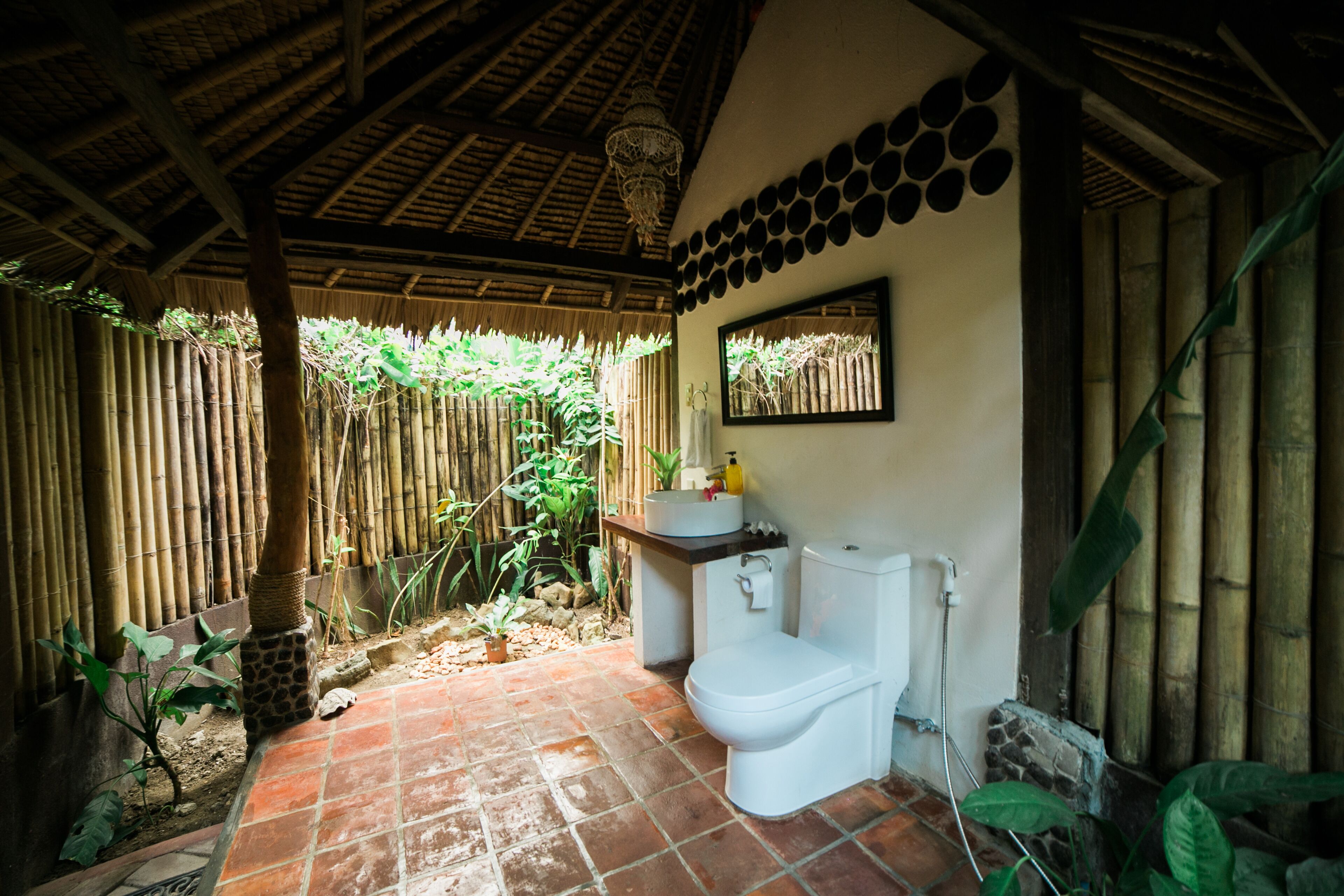 Une salle de bain avec des toilettes, un lavabo et un miroir. À l’extérieur, on trouve une clôture en bambou et le sol est carrelé.