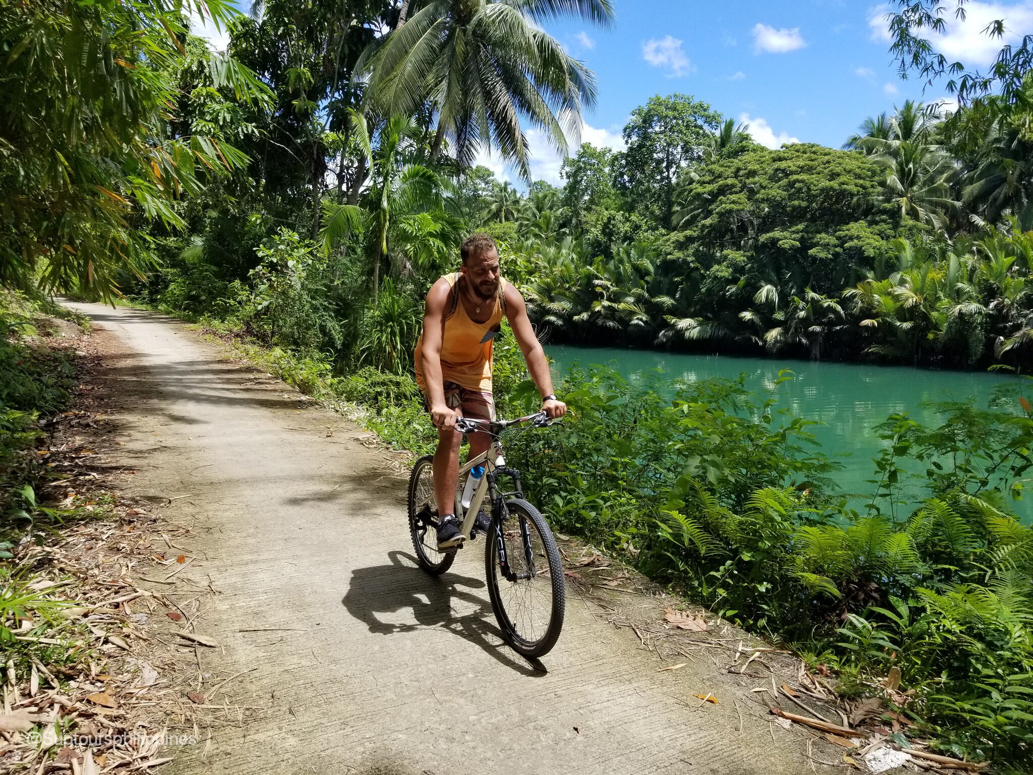 A person riding a bicycle on a concrete path surrounded by lush greenery and a body of water.