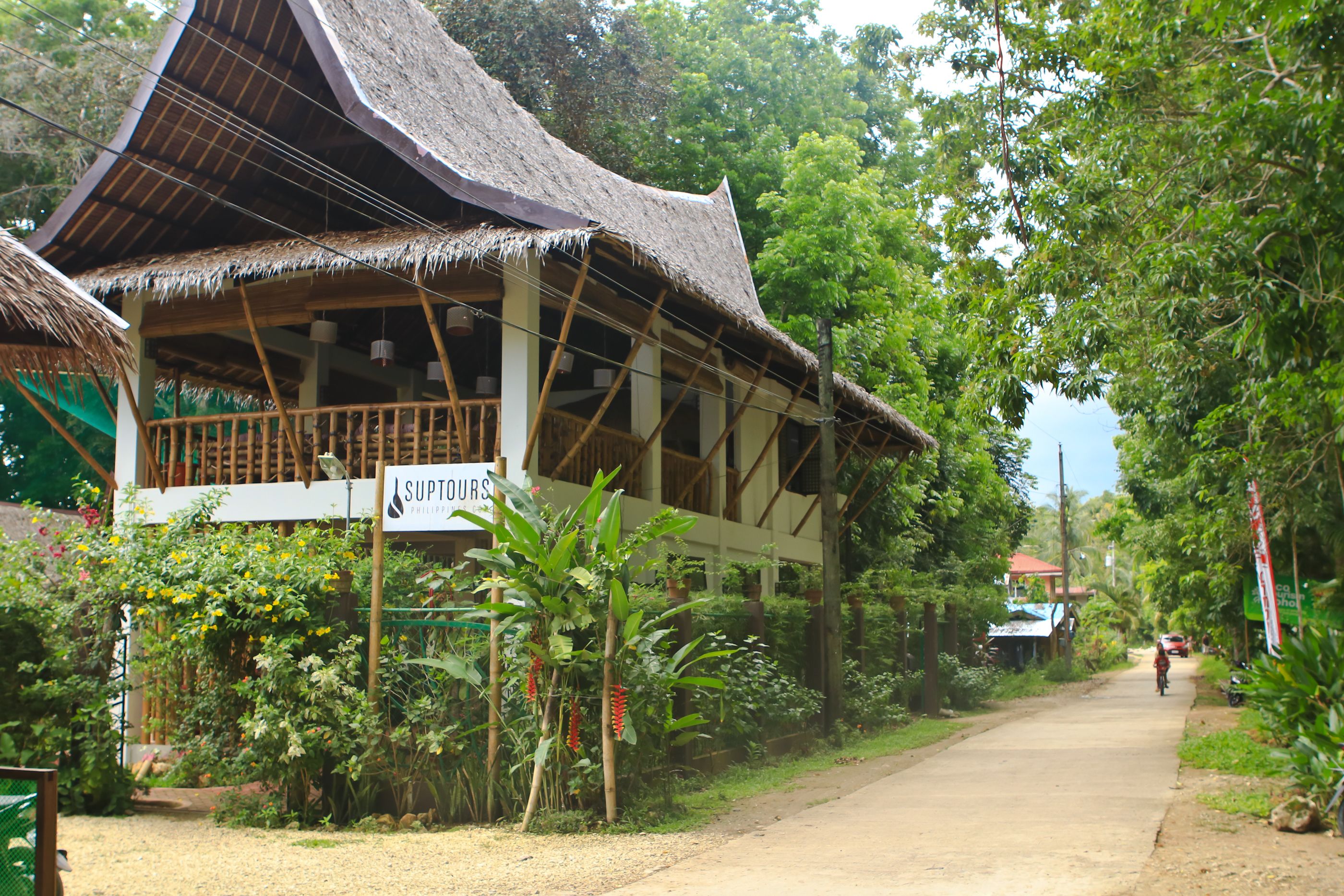 A traditional wooden building with a thatched roof, surrounded by lush greenery and a dirt road.