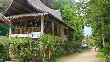 A traditional wooden building with a thatched roof, surrounded by lush greenery and a dirt road.