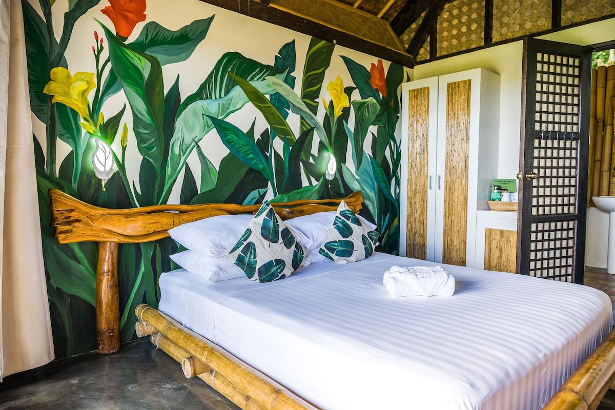Una cama con cabecera de madera, ropa de cama blanca y almohadas con estampado de hojas tropicales.
