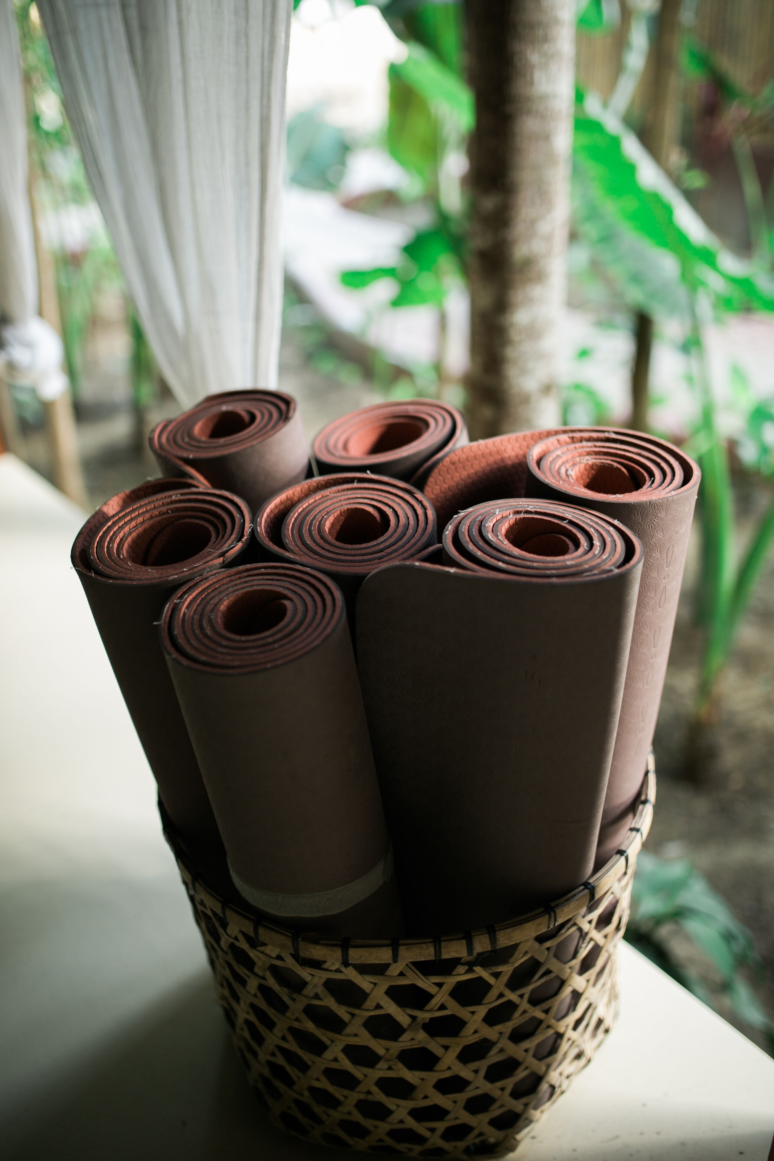 A woven basket containing several rolled-up brown mats, placed on a table with a blurred background of greenery.