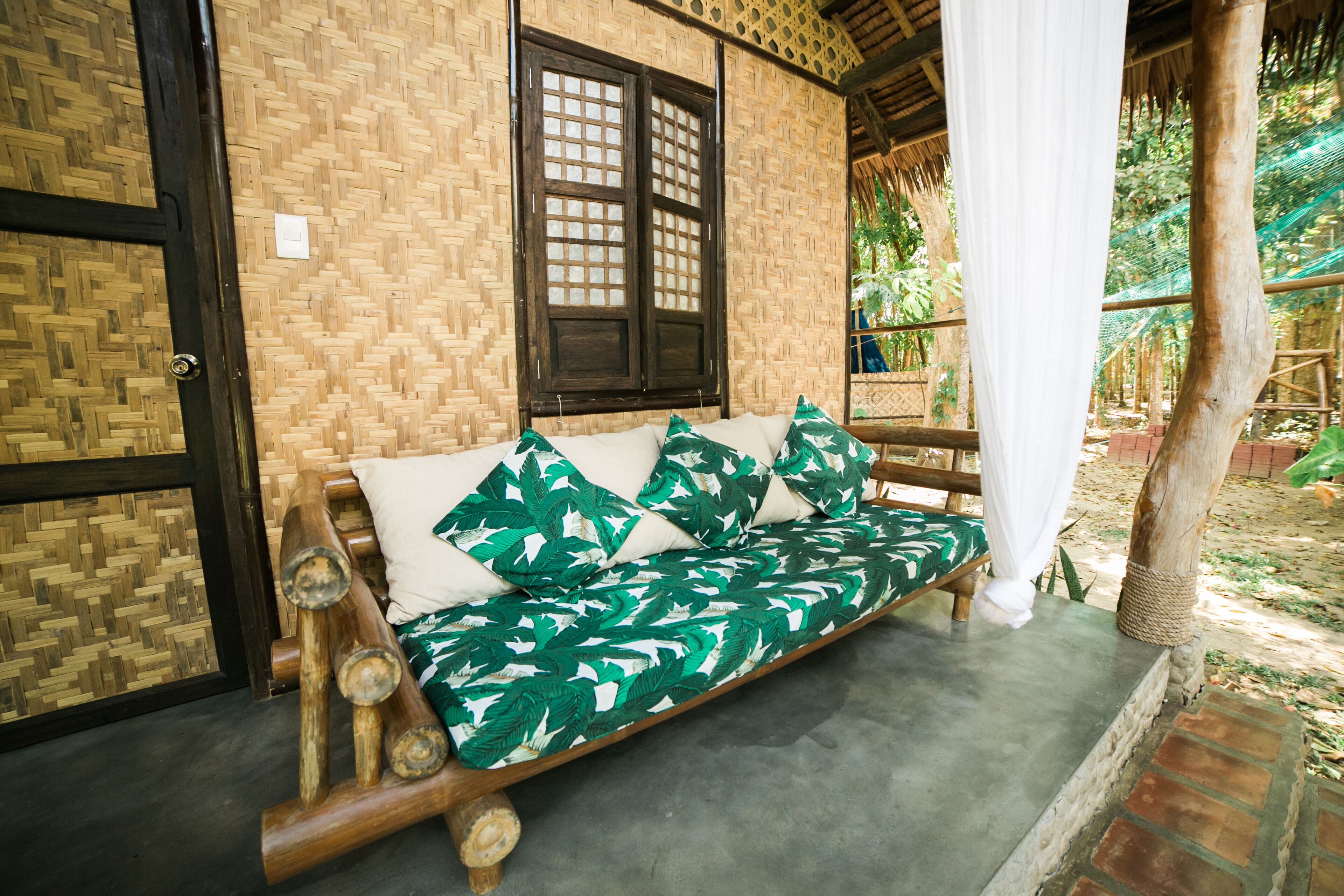 A wooden sofa with a green leaf pattern, positioned in front of a woven wall and a wooden door.