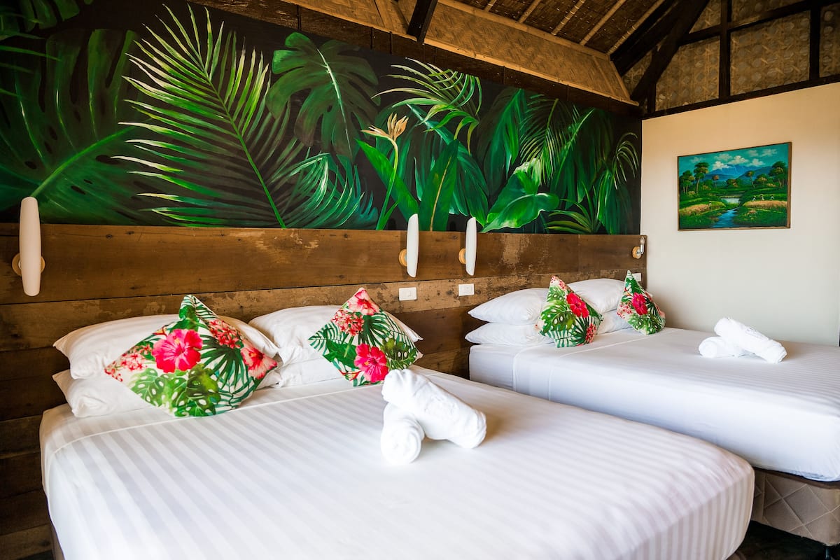 Dos camas con sábanas blancas, almohadas florales y toallas. Un gran mural tropical y un cuadro de paisaje decoran las paredes.