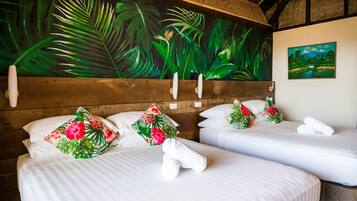 Dos camas con sábanas blancas, almohadas florales y toallas. Un gran mural tropical y un cuadro de paisaje decoran las paredes.