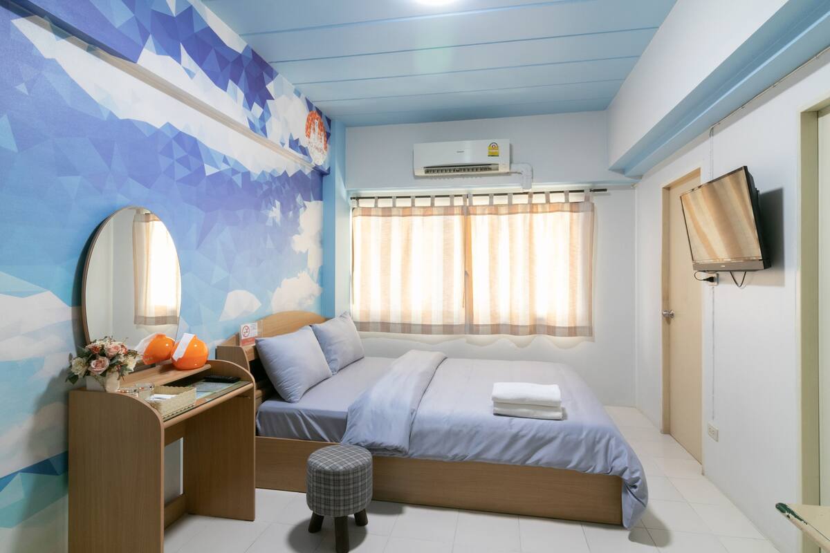 A bedroom with a bed, a desk with a chair, a mirror, a window with curtains, an air conditioning unit, and a television on the wall.