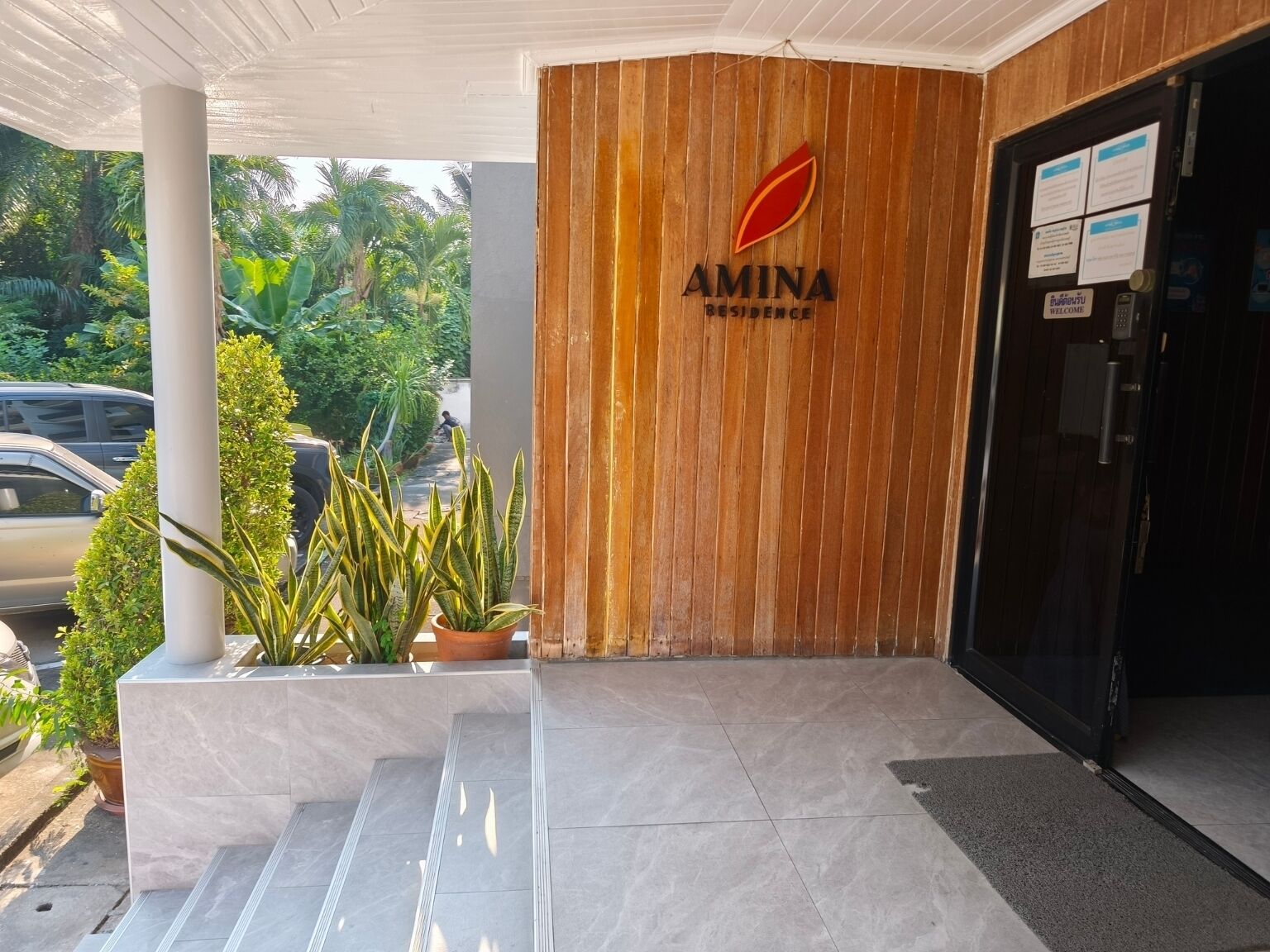A hotel entrance with a wooden facade, a sign reading "AMINA RESIDENCE," and a black door with a welcome sign.