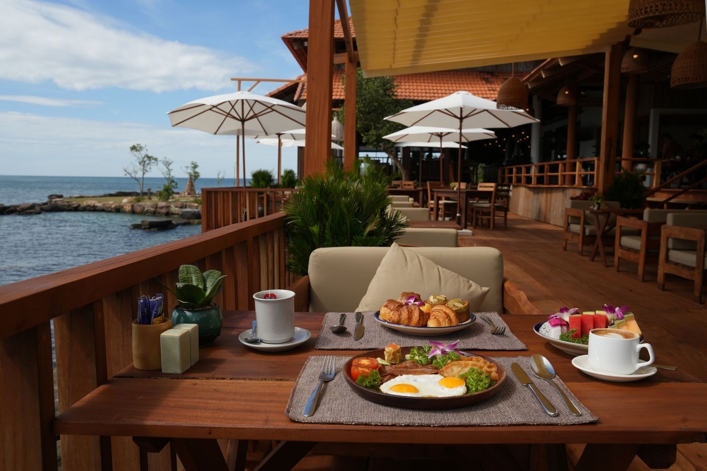 A wooden deck with outdoor dining, a view of the sea, and umbrellas providing shade.