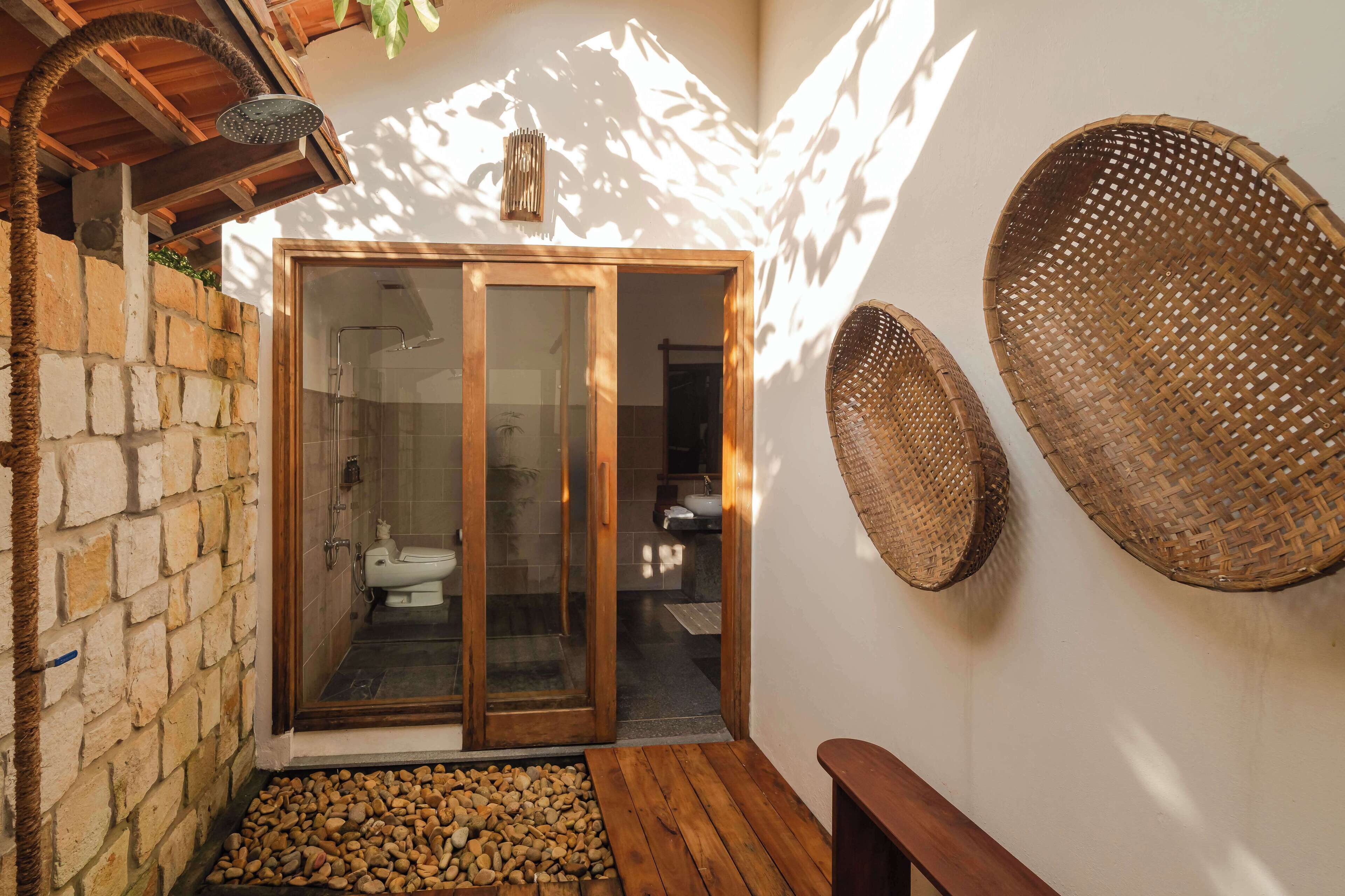 A bathroom with a glass door, a shower, and a toilet. There are woven baskets on the wall and a wooden bench.