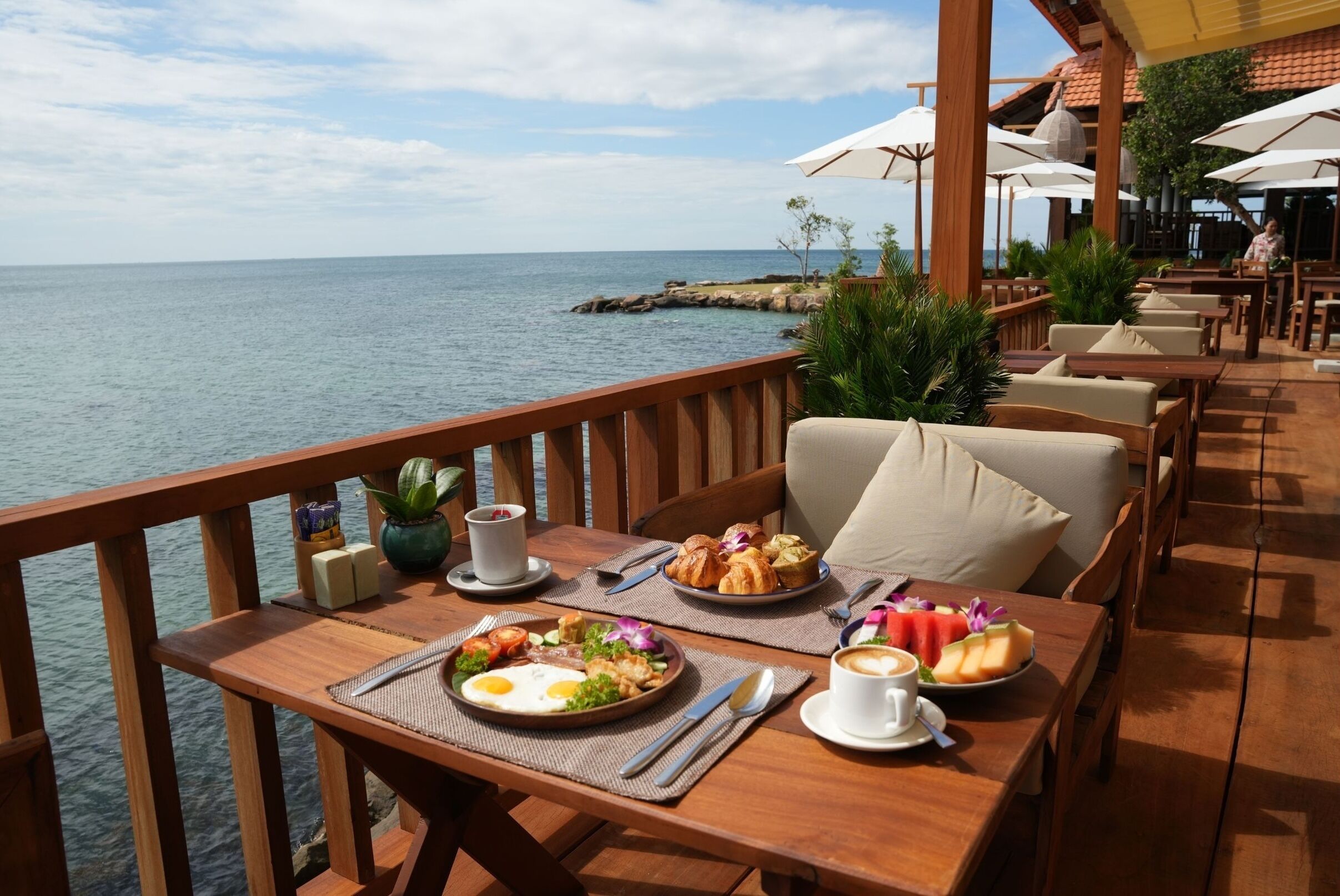A wooden deck with tables and chairs overlooking the sea, featuring breakfast items, a cup of coffee, and a view of the ocean.