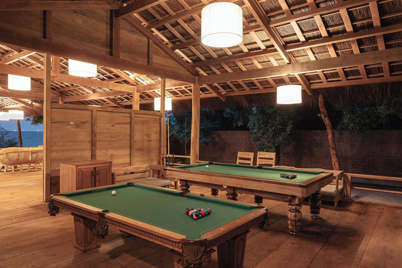 An indoor pool area with two pool tables, wooden furniture, and a ceiling with exposed beams and hanging lights.