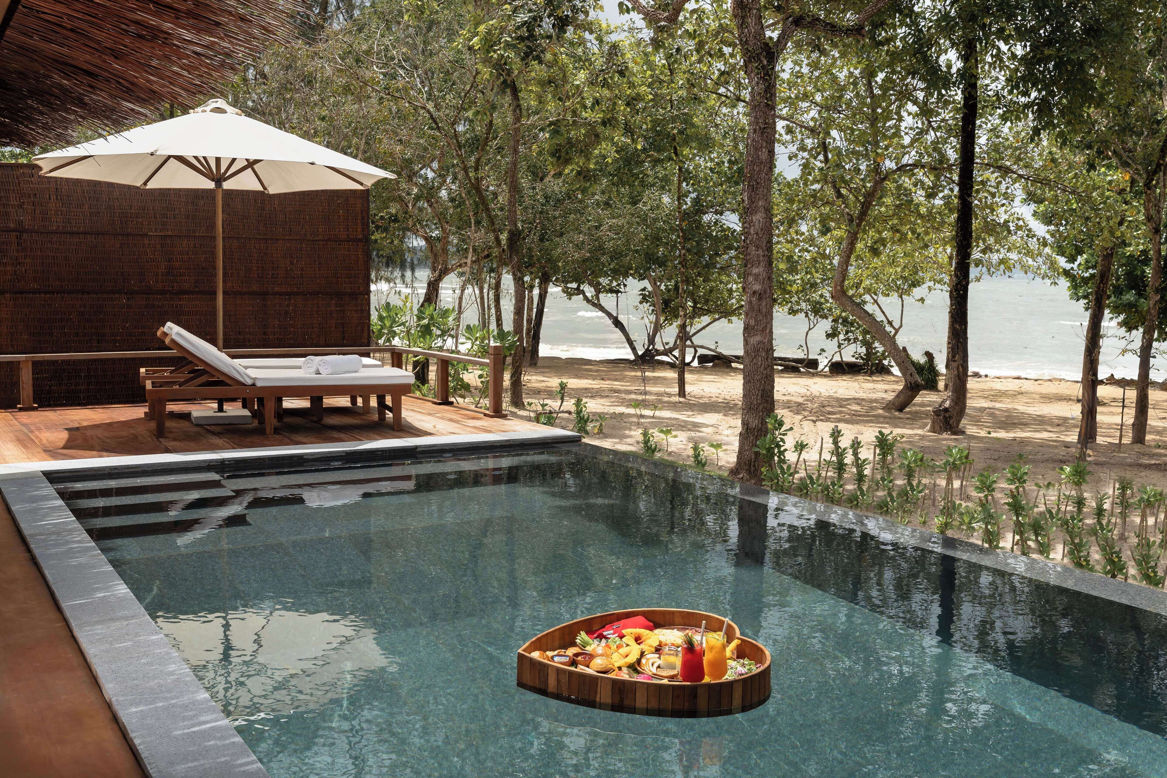A swimming pool with a floating tray of food, lounge chairs, and a beach view.