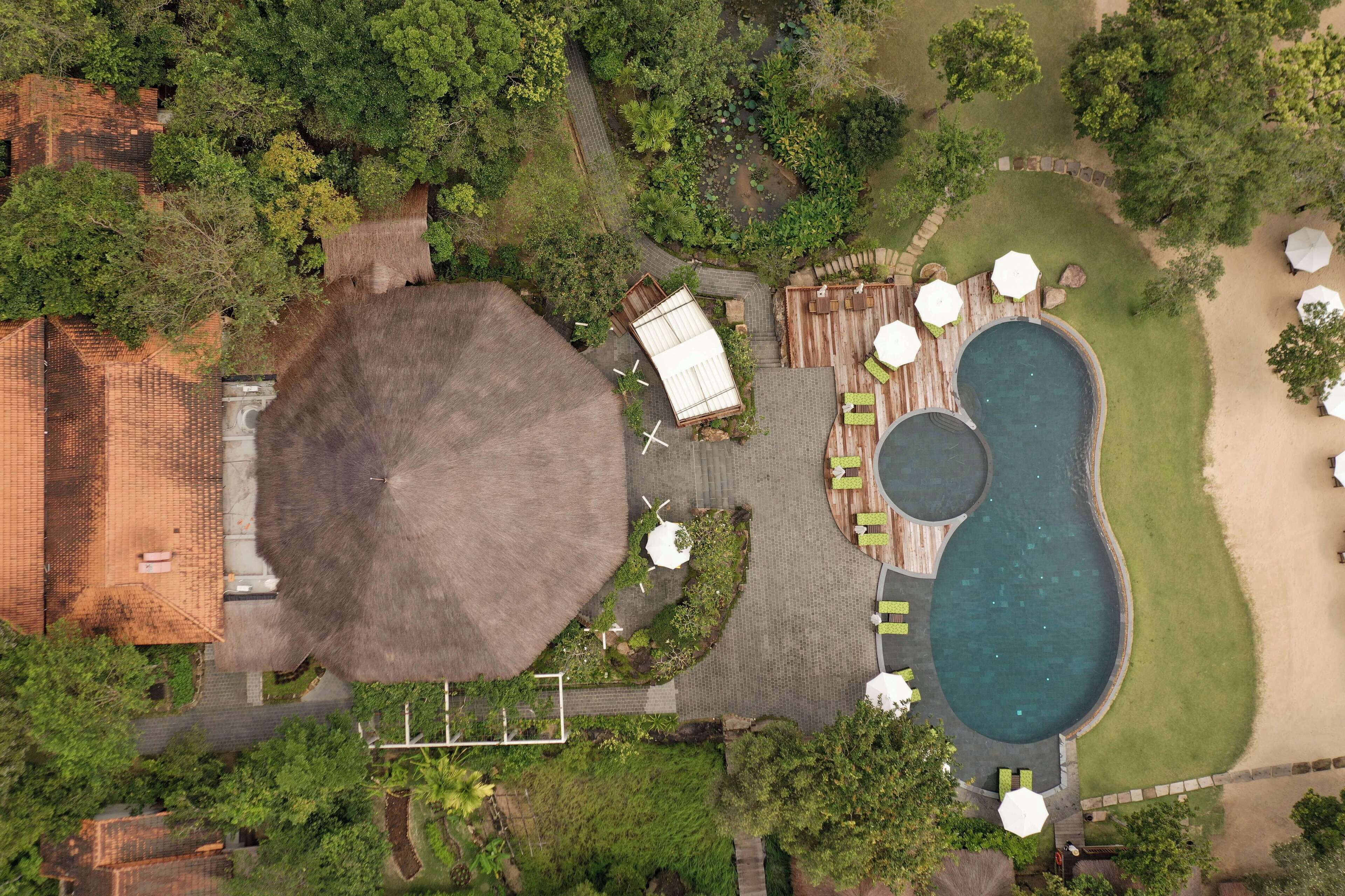 An aerial view of a resort with a large pool, a thatched-roof structure, and a well-maintained garden.