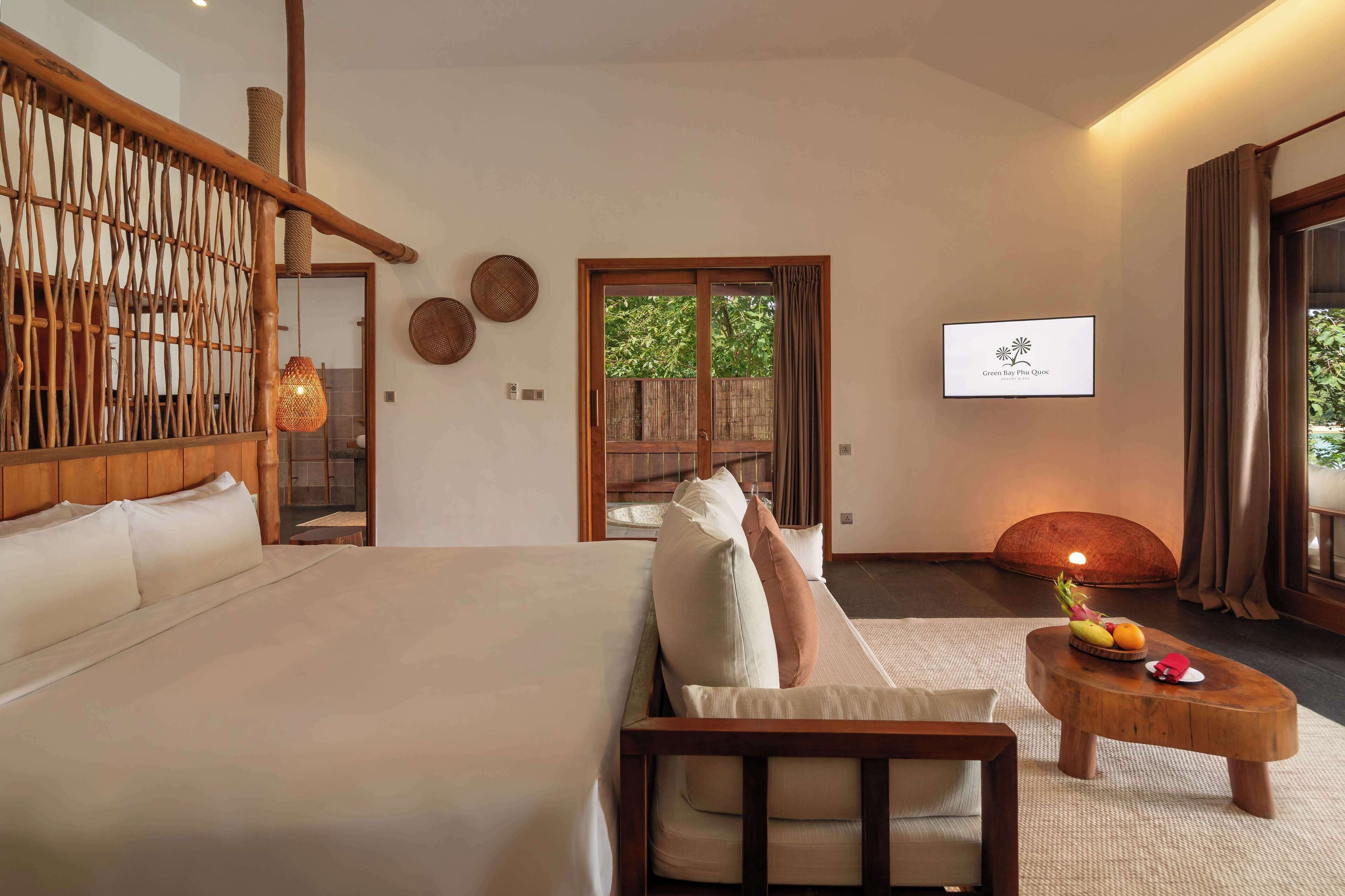 A modern hotel room with a large bed, a wooden headboard, a small sofa, a wooden coffee table with fruit, and a TV on the wall.