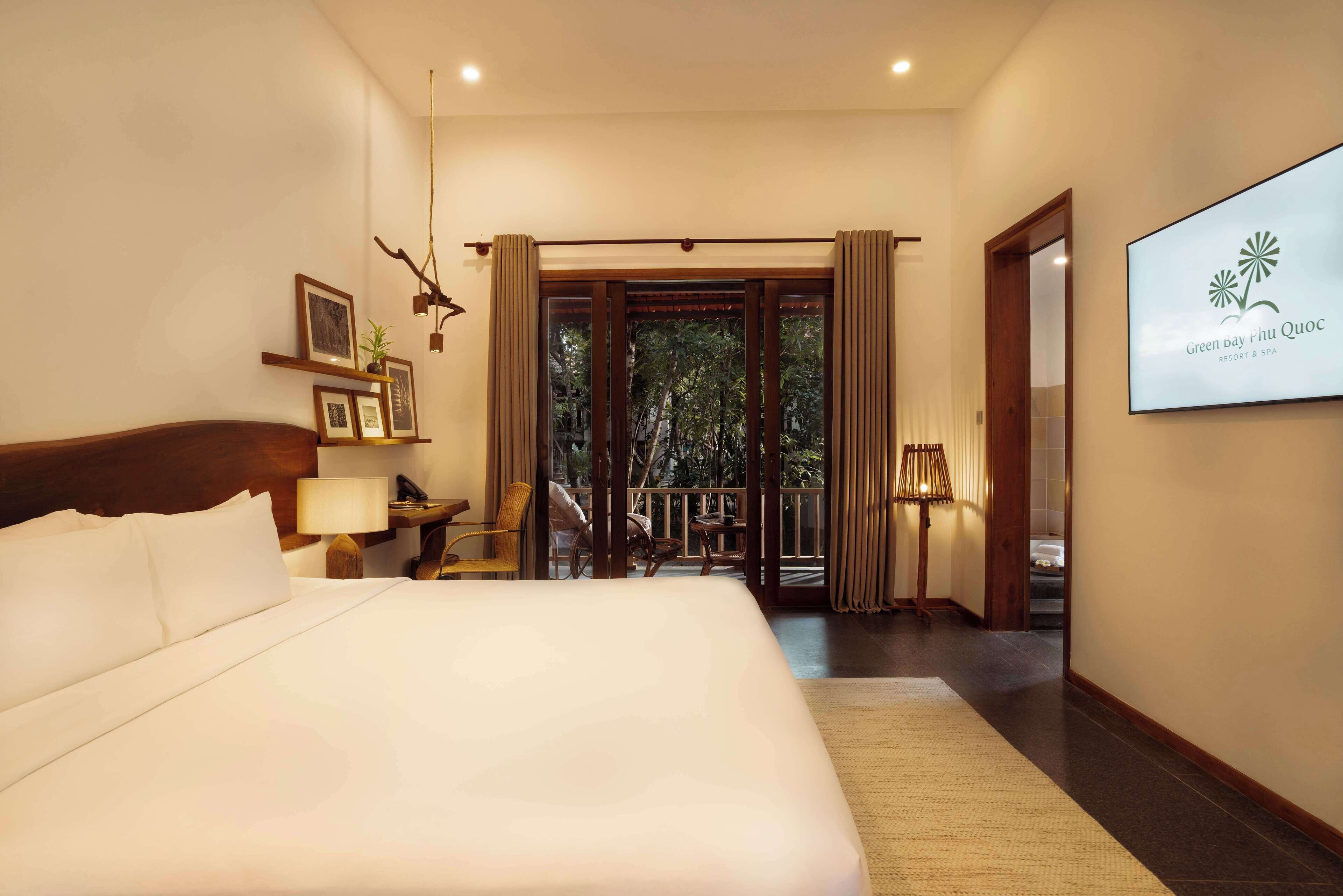 A hotel room with a large bed, a desk with a lamp, a chair, a view of the outdoors through a glass door, and a sign for Green Bay Phu Quoc Resort & Spa.