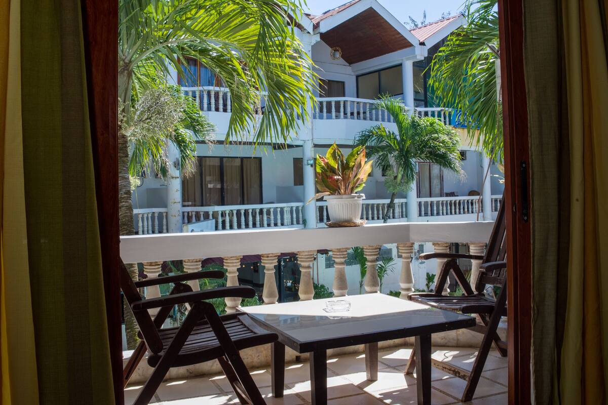 An outdoor balcony with a table and chairs, surrounded by potted plants and a building with balconies.