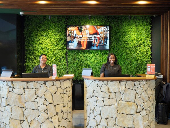 Two receptionists at a reception desk with a green wall and a television above.