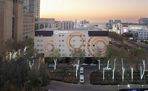 Un edificio de hotel con un gran logo circular, varios pisos y una zona de estacionamiento con árboles.