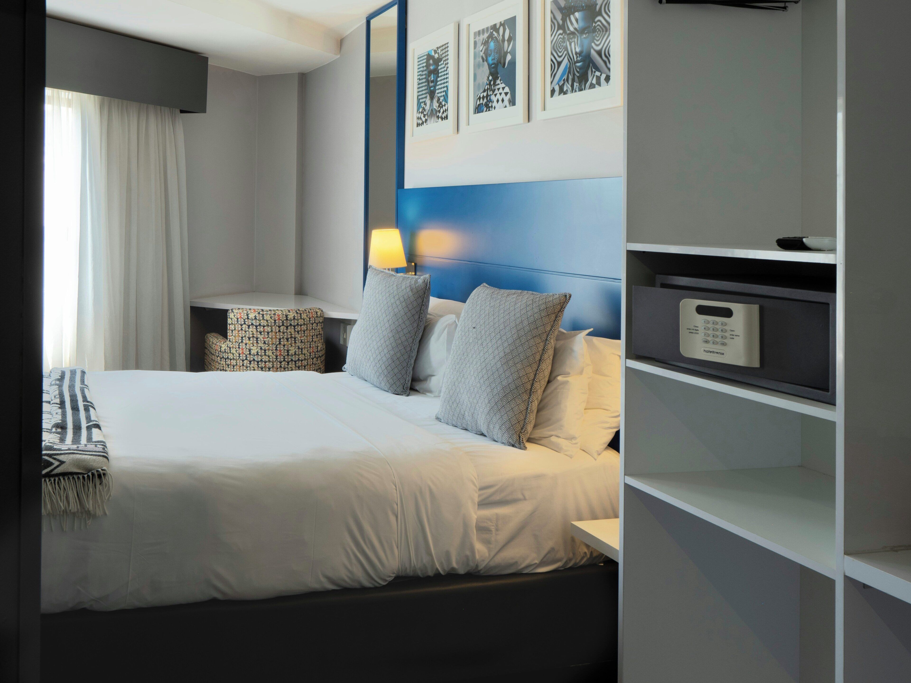 A hotel room with a bed, bedside tables, a lamp, a nightstand, a shelf with a remote control, and framed artwork on the wall.