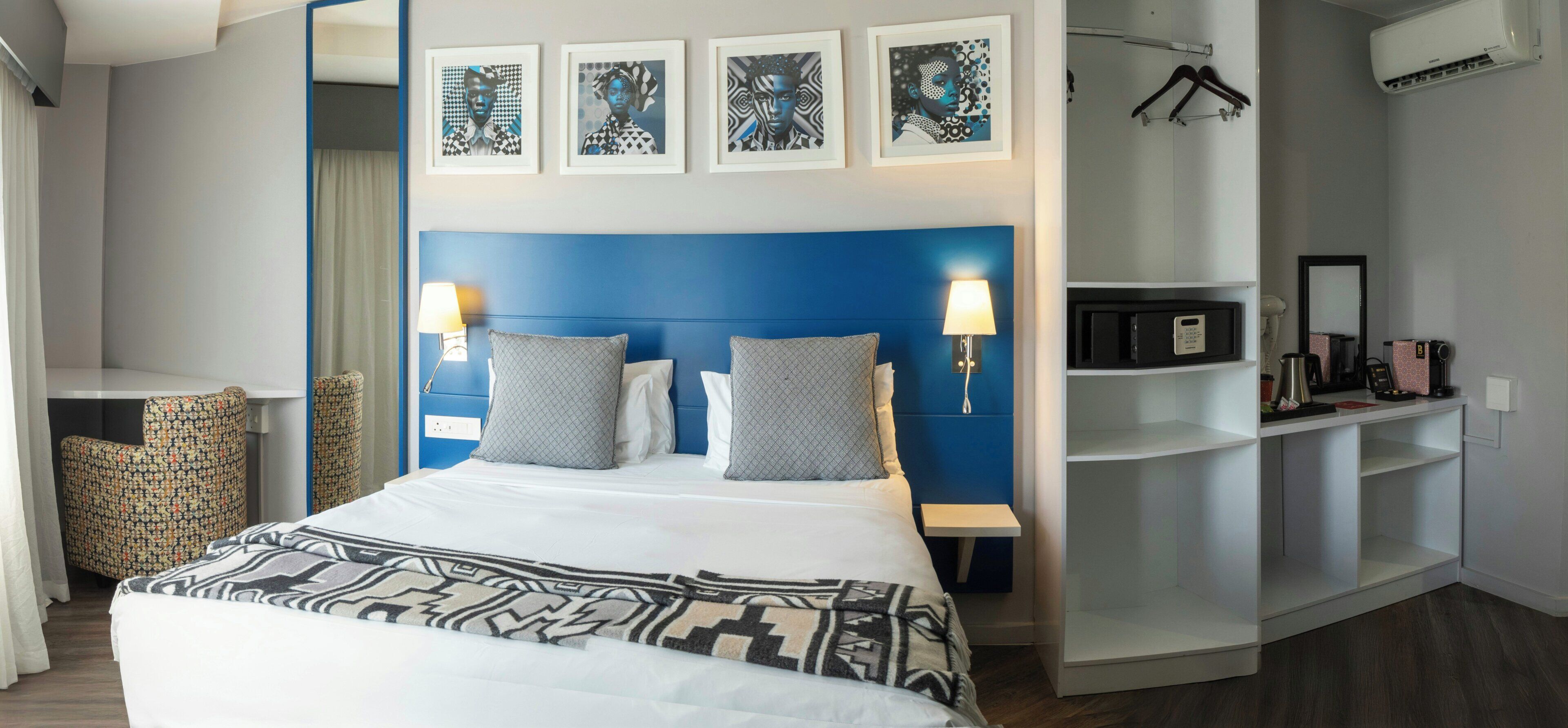 A hotel room with a bed, two bedside lamps, a headboard, a wardrobe, a desk, a chair, and a mirror.