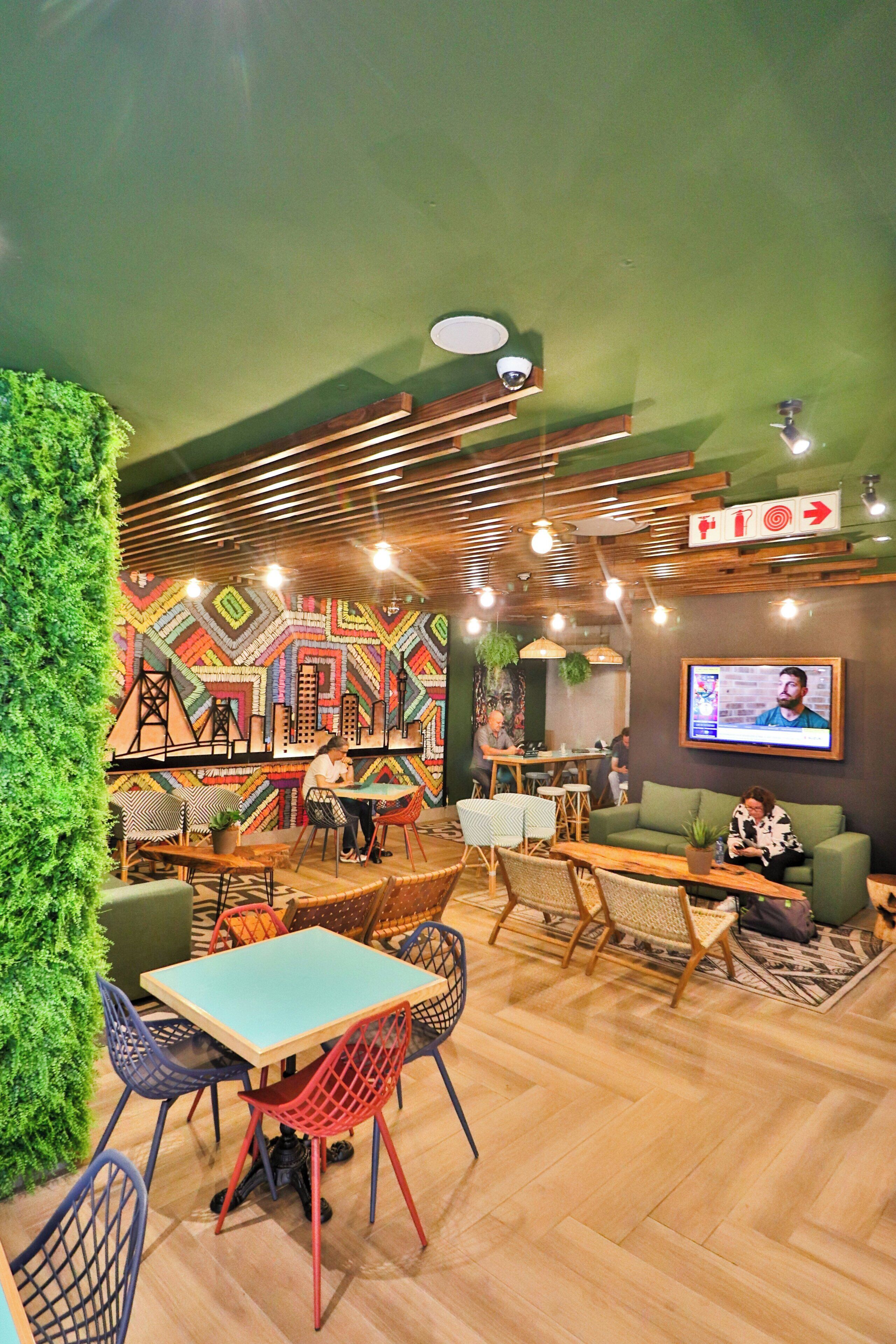 Un interior moderno con una pared verde, techo de madera y un vibrante cuadro de arte geométrico. Hay mesas y sillas, un televisor y un sofá verde.
