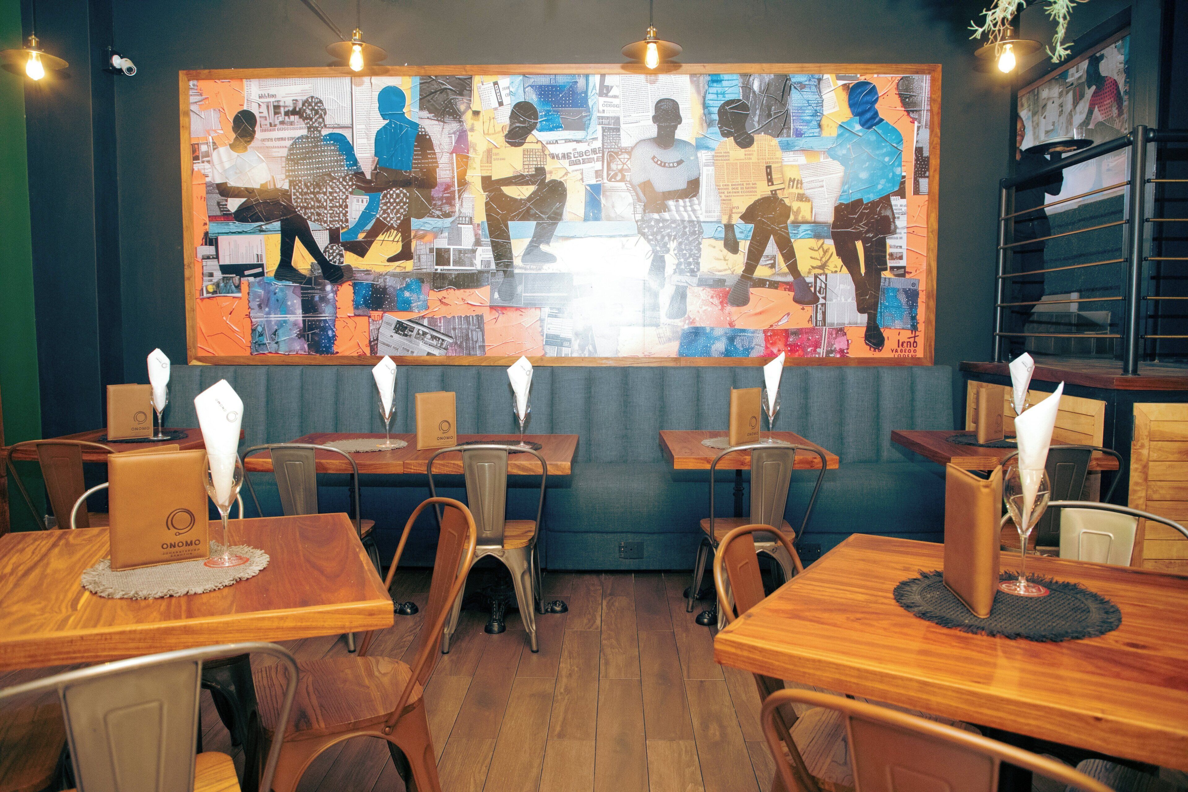 A restaurant interior with wooden tables, chairs, and a large abstract mural on the wall.