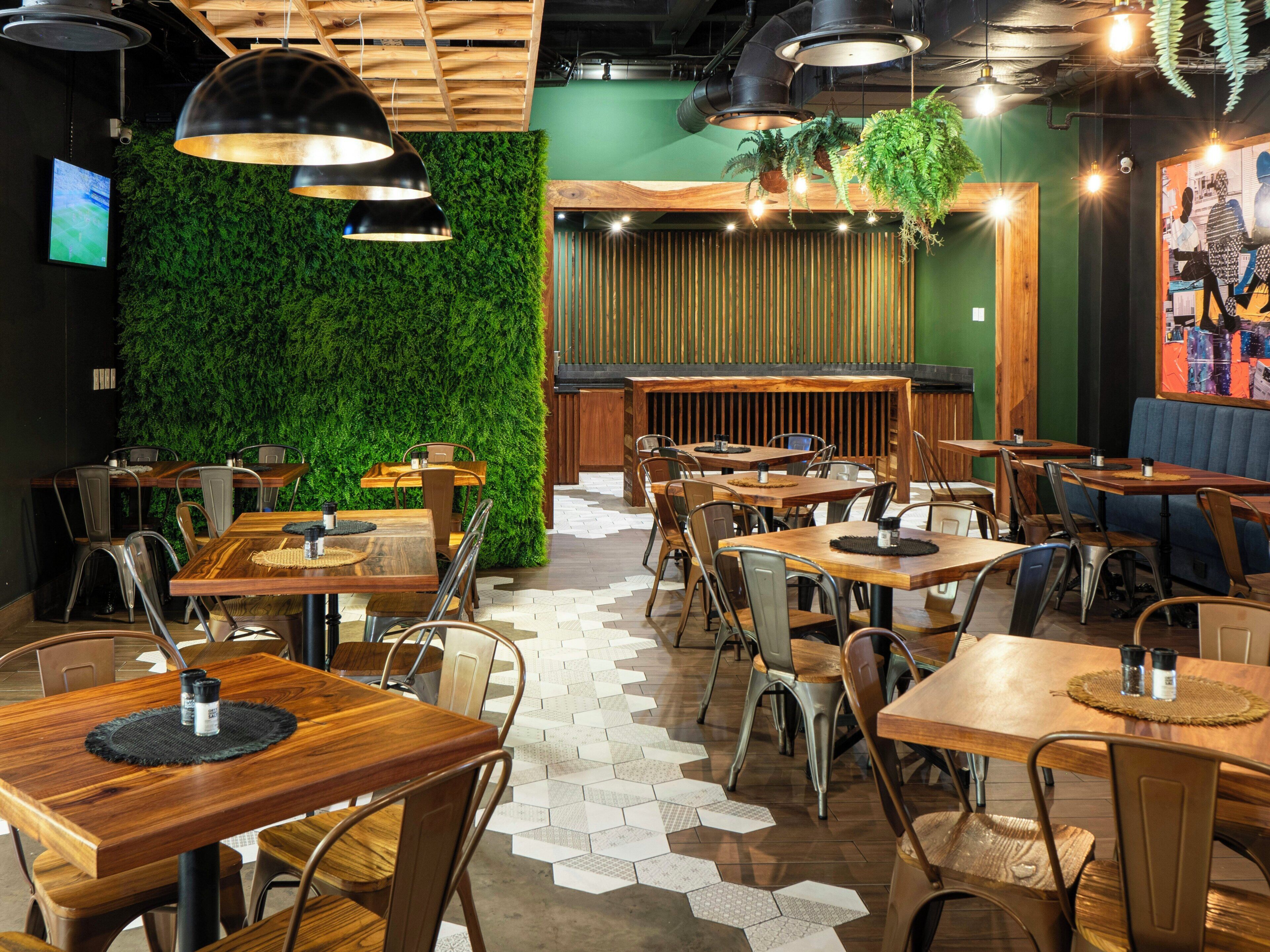 A restaurant interior with wooden tables and chairs, a green wall with plants, and pendant lighting.