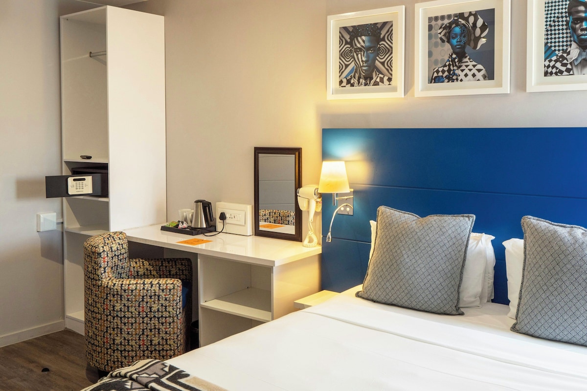 Quarto de hotel com cama, escrivaninha, cadeira, espelho e obras de arte na parede.