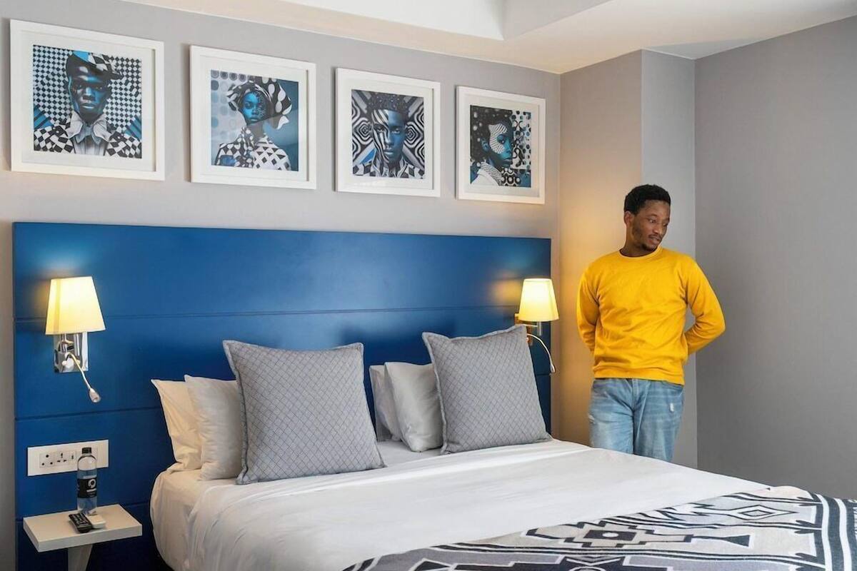 Habitación de hotel con cabecera azul, cama con ropa de cama blanca, dos lámparas de noche y cuatro cuadros enmarcados en la pared.