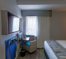 Una camera d'albergo con un letto, una scrivania, una sedia, una televisione e una finestra con tende.