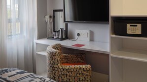 A hotel room with a bed, a TV mounted on the wall, a hairdryer, and a coffee maker on a shelf. - ONOMO Hotel Johannesburg Sandton (Sandton)