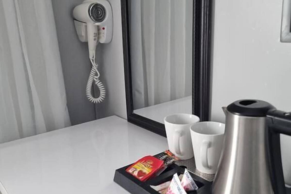 Une chambre d’hôtel avec un comptoir blanc, un sèche-cheveux, un miroir, deux tasses blanches, une cafetière en argent et un plateau contenant des sachets de thé et un paquet rouge.
