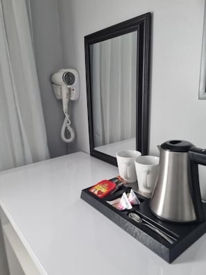 Une chambre d’hôtel avec un comptoir blanc, un sèche-cheveux, un miroir, deux tasses blanches, une cafetière en argent et un plateau contenant des sachets de thé et un paquet rouge.