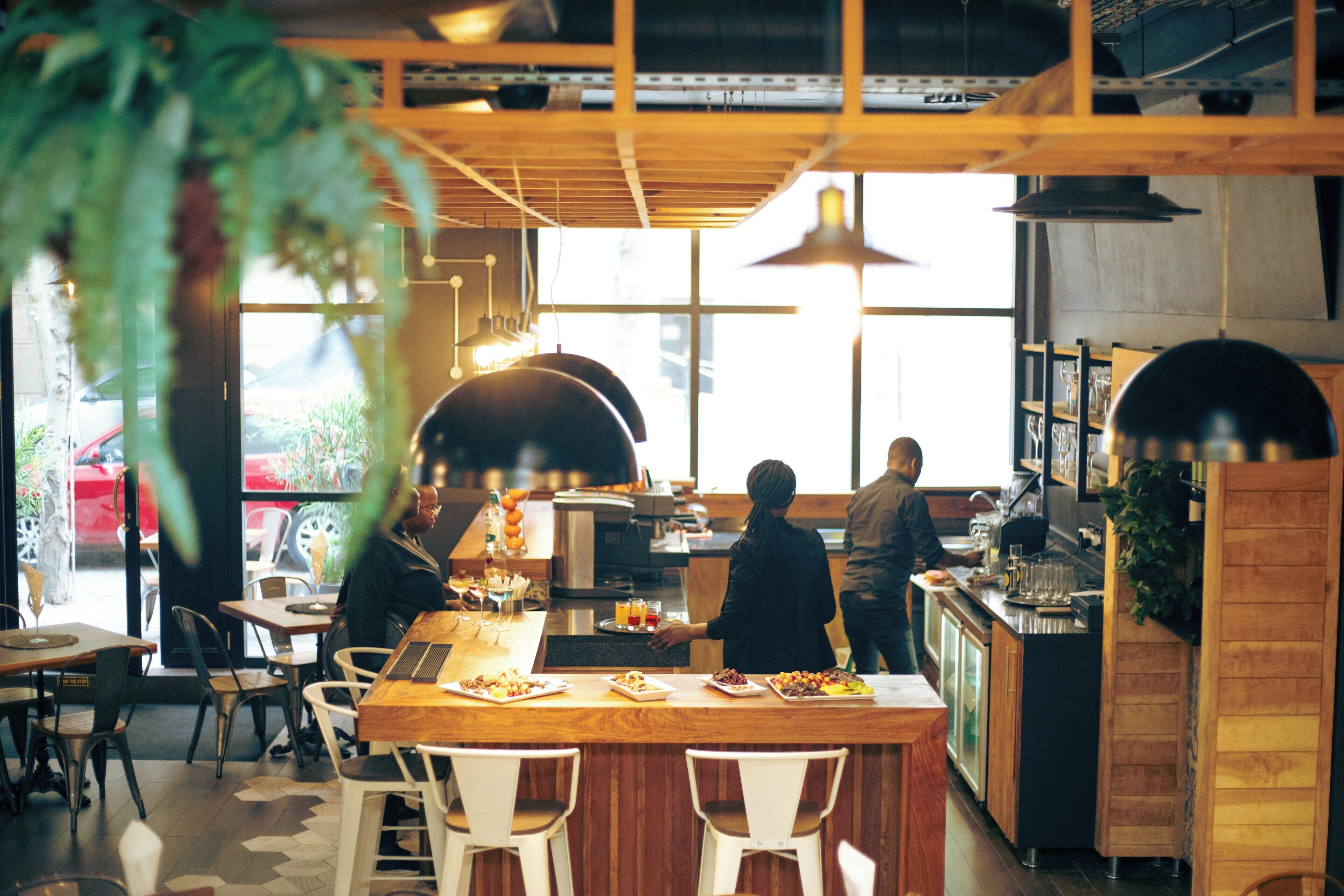 An indoor dining area with wooden tables and chairs, a counter with a person preparing food, and large windows allowing natural light.