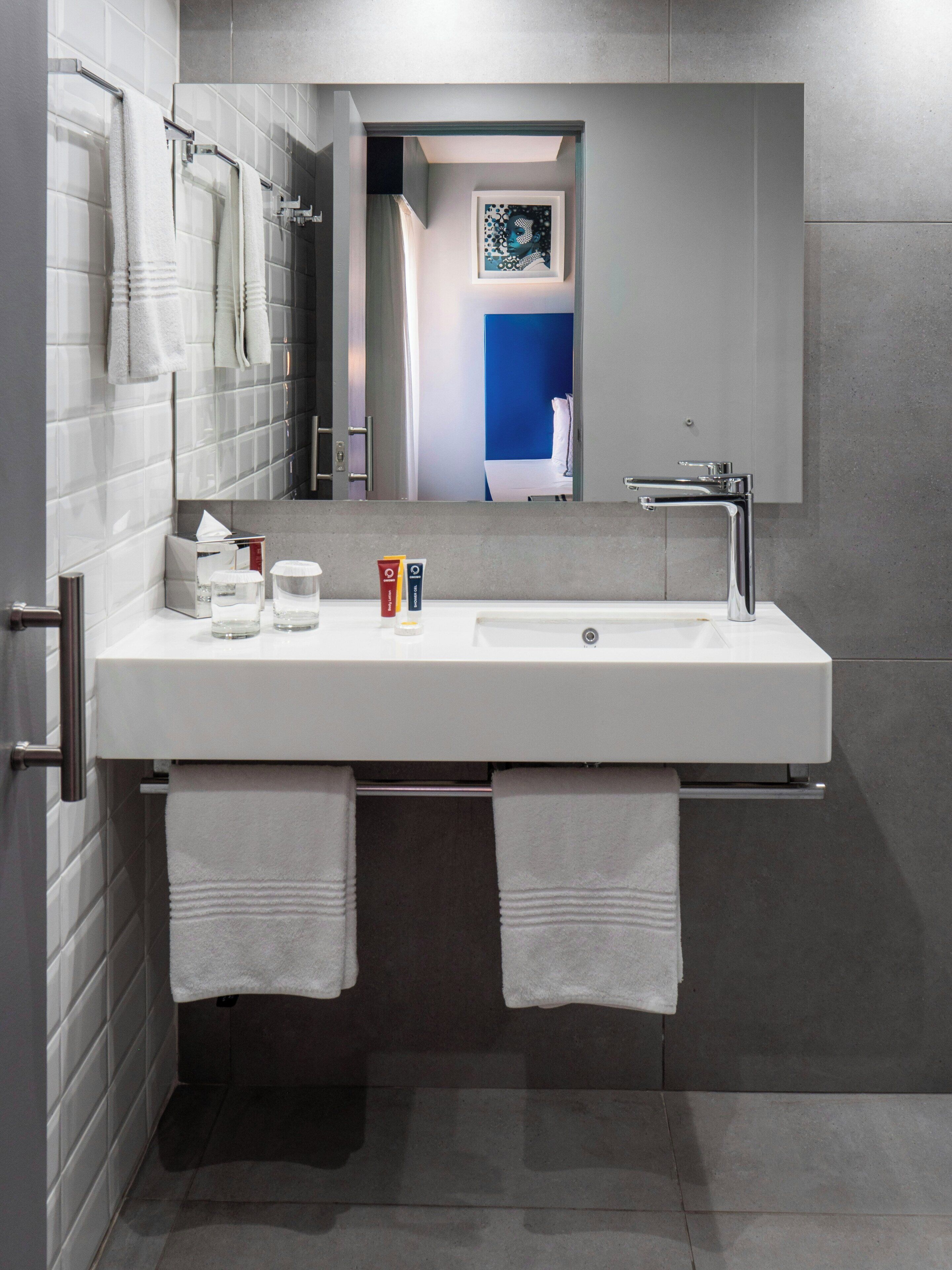 A modern bathroom with a white sink, two towels, a mirror, and a framed artwork on the wall.