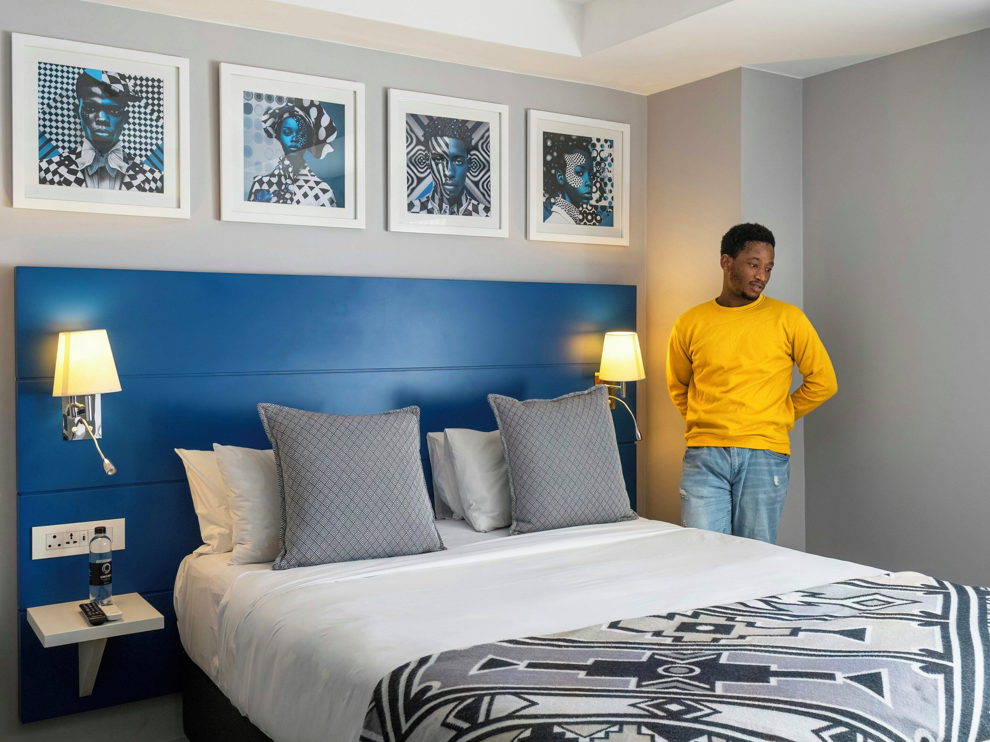 A hotel room with a blue headboard, a bed with patterned pillows, bedside lamps, and artwork on the wall.