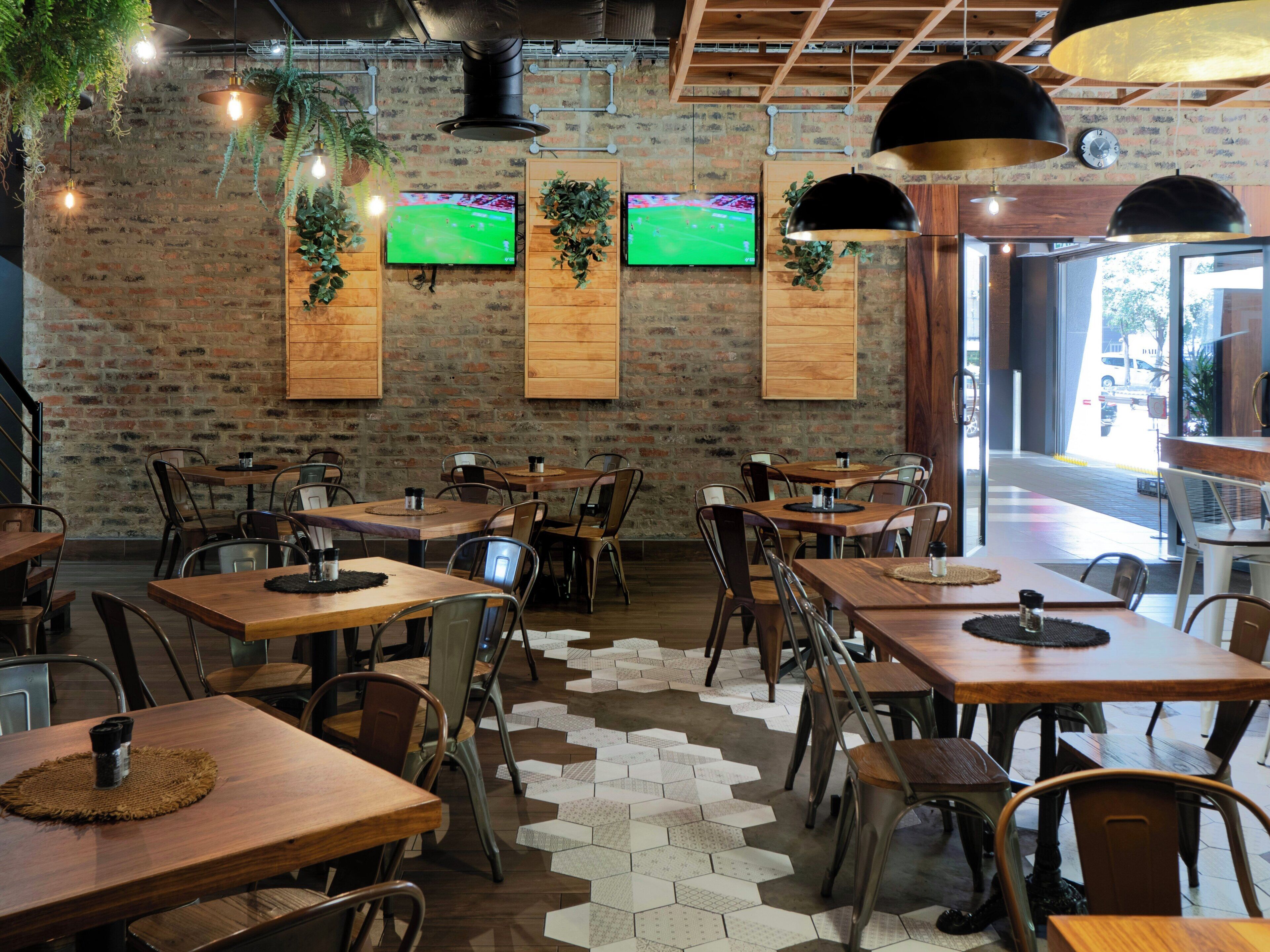 Interior de restaurante con paredes de ladrillo, mesas de madera, sillas de metal, plantas colgantes y televisores instalados.