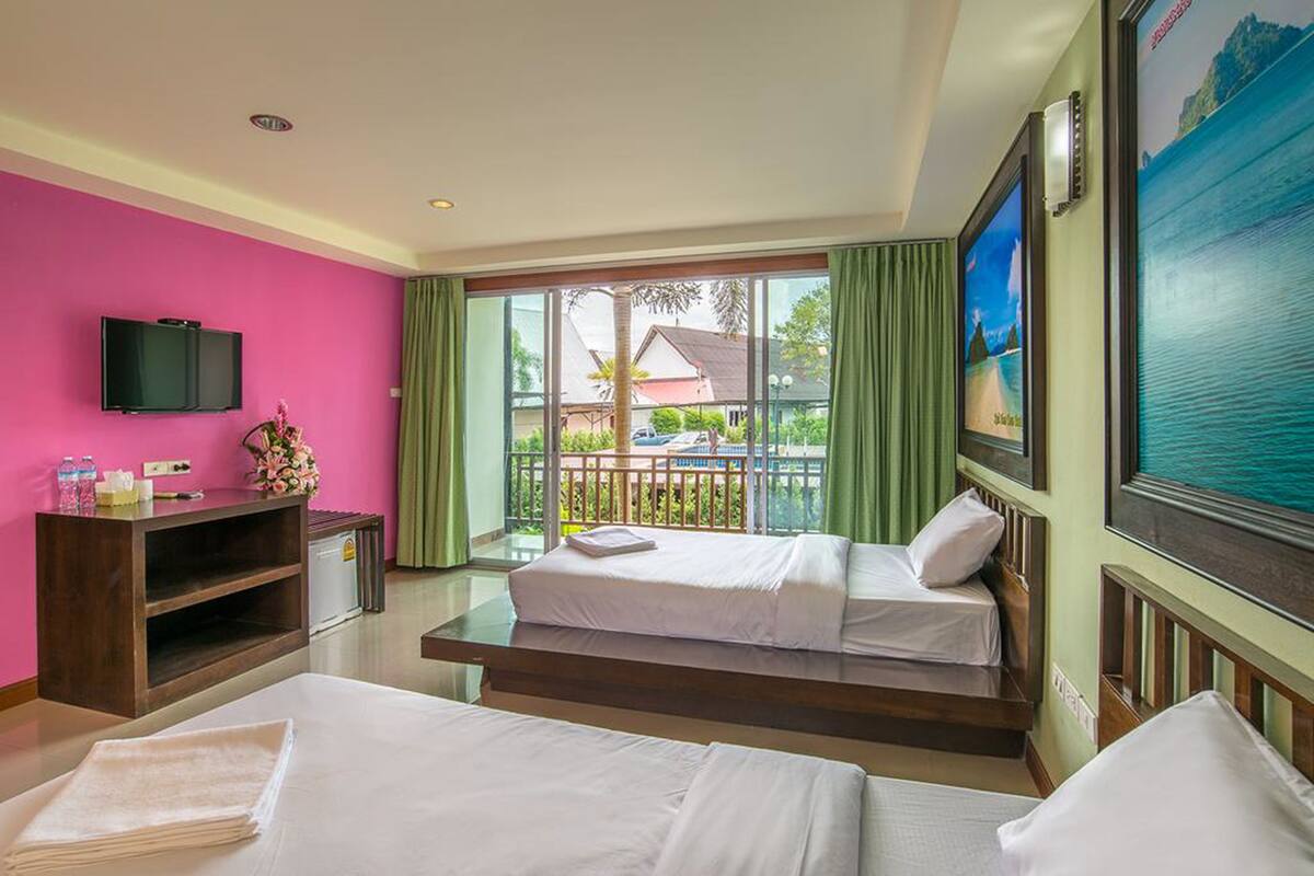 Une chambre d’hôtel avec deux lits, une télévision, un bureau avec un ordinateur et un balcon donnant sur une maison et un palmier.