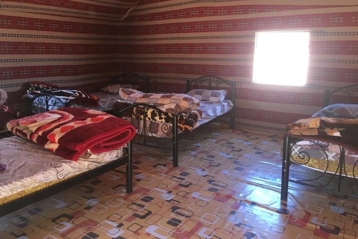 A room with three beds, each with a patterned bedspread, and a window providing natural light. The floor has a geometric patterned design.