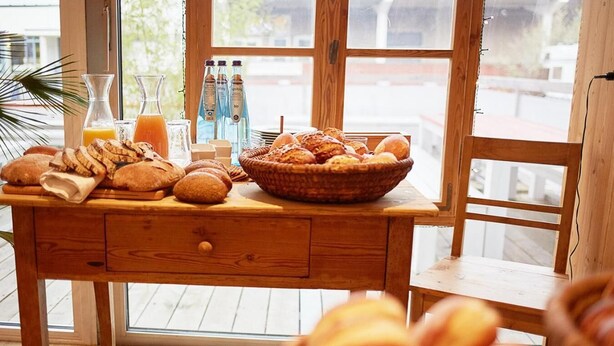 A wooden table with various breads and pastries, a glass of orange juice, and bottles of water. - Hotel Capima (Cologne)